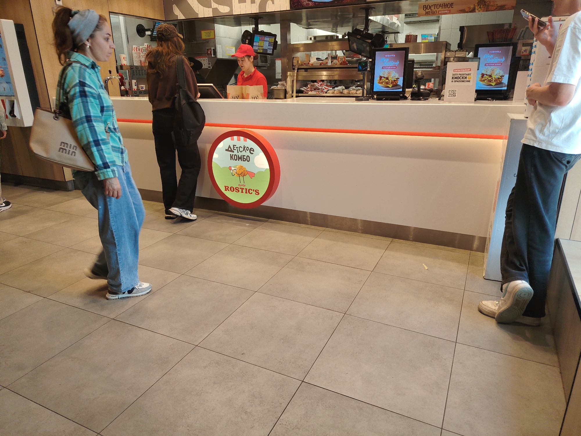 In Yekaterinburg, Russia, a scene unfolds within a fast-food restaurant, likely Rostic's, as evidenced by a circular logo featuring a cartoon chicken and the text "ROSTIC'S" prominently displayed on the counter. The environment is characterized by tiled flooring and a white counter area.
At the counter, three individuals are visible. To the left, a woman with a light blue and turquoise plaid shirt and light blue jeans stands facing away from the camera, carrying a beige handbag. She appears to be looking towards the counter. To her right, another woman, wearing a brown hooded jacket and dark pants, is also at the counter, partially obscured. Behind the counter, a female employee in a red shirt and a red cap is actively serving customers, holding a paper bag. Two digital menu screens display images of burgers, and a QR code is visible on a white sign. On the far right, a person wearing a white t-shirt and dark pants stands holding a smartphone. The overall atmosphere suggests a typical day at a fast-food establishment.