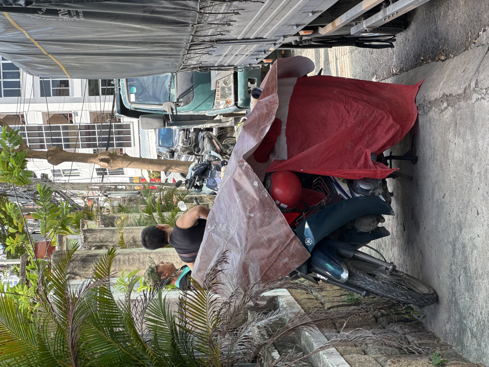 The image depicts a narrow street in Nha Trang, Vietnam, with a focus on a parked motorcycle covered by a red tarp. Two men are standing near the motorcycle, with one wearing a camouflage hat and the other a black sleeveless shirt. In the background, a green truck and a larger truck with a black tarp are parked, indicating a busy street scene. The environment includes palm trees, a stone wall, and a building with multiple windows. The overall impression is of a typical urban setting with everyday activities.