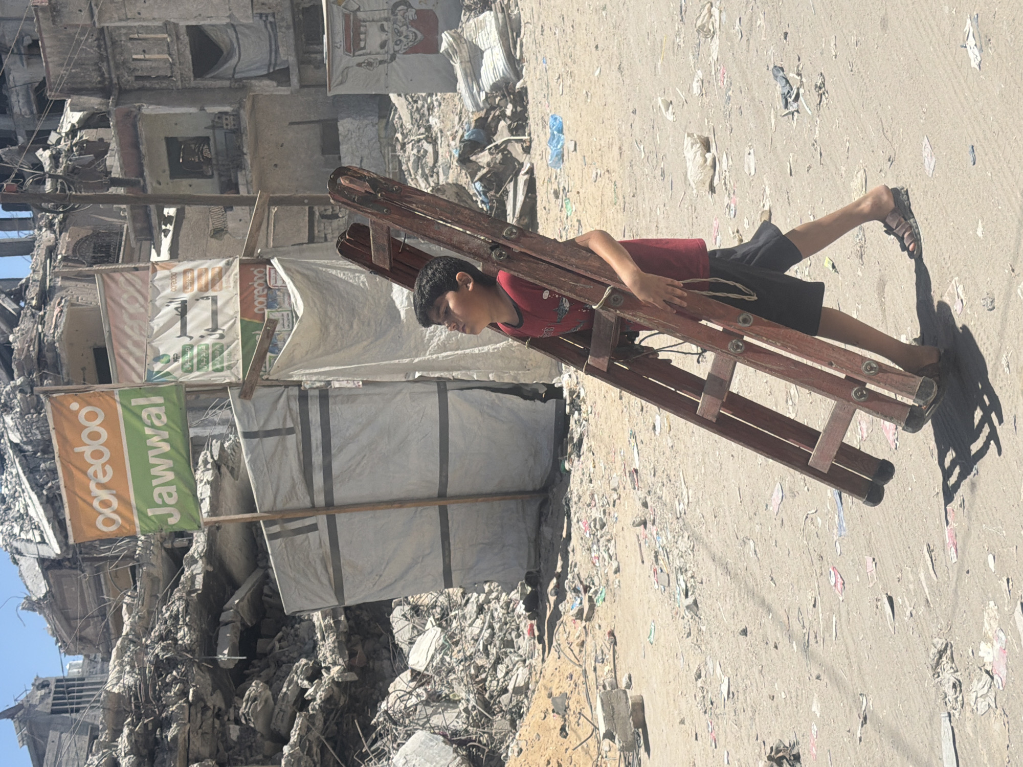 A young boy in a red t-shirt and dark shorts walks in the street of Gaza City, Palestinian Territories, carrying a wooden ladder. The street is littered with debris, including concrete rubble, plastic bags, and paper scraps, indicating destruction. Behind the boy, buildings are heavily damaged, with collapsed structures and exposed rebar, suggesting recent conflict. Two large banners are visible in the background: one with the Ooredoo and Jawwal logos in orange and green, and another, partially obscured, with Arabic text and the Ooredoo logo. The sky is clear and blue, and the lighting suggests it is daytime. The overall scene depicts a post-conflict environment with signs of both ongoing life and widespread devastation.