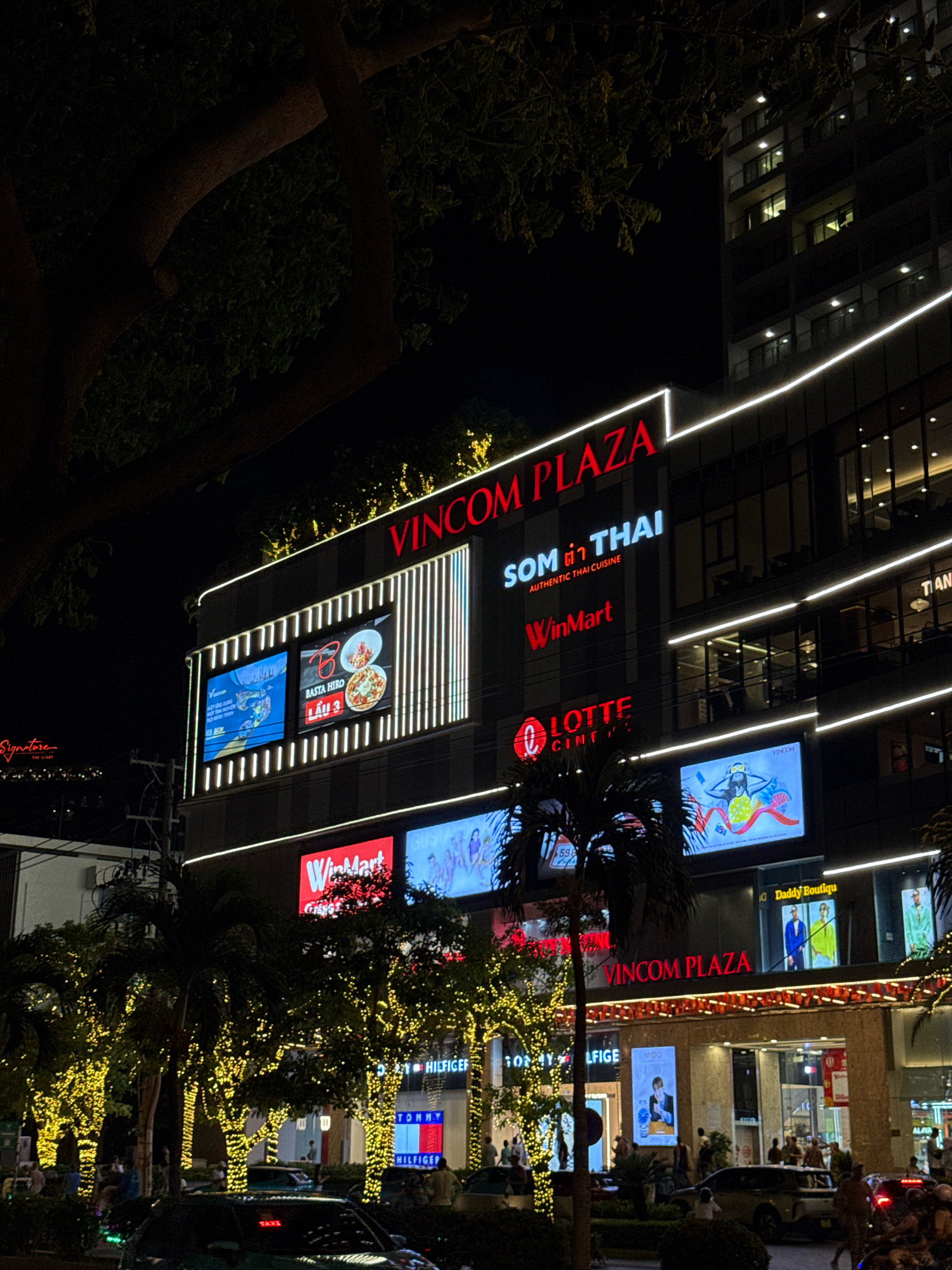 The image depicts a nighttime view of Vincom Plaza in Nha Trang, Vietnam, as evidenced by the prominent "VINCOM PLAZA" signage. The multi-story building is illuminated with various signs and lights, highlighting its commercial purpose. Large digital billboards display advertisements, including one for "SOM THAI AUTHENTIC THAI CUISINE" and another for "LOTTE CINEMA." The lower levels of the building feature storefronts for brands like "WinMart" and "Tommy Hilfiger," indicated by their respective logos. The surrounding environment is decorated with numerous strings of yellow fairy lights draped over trees and other structures, creating a festive atmosphere. Several palm trees are visible in the foreground. Pedestrians are scattered throughout the scene, some walking, others appearing to be socializing. Vehicles, including cars and motorcycles, are present on the street in front of the plaza. The architecture suggests a modern urban setting.