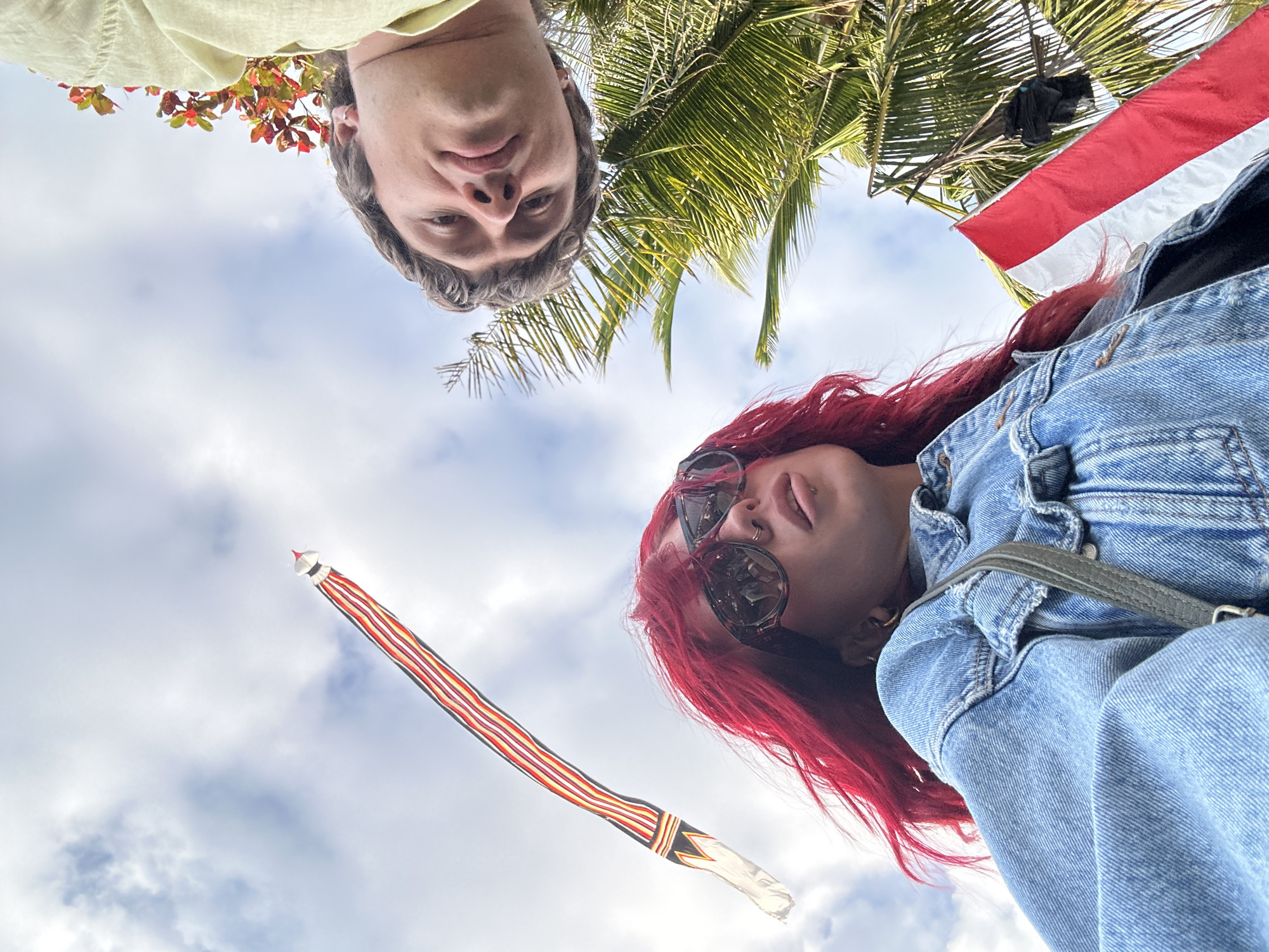 In the city of Mengwi, Indonesia, a low-angle shot captures two individuals against a backdrop of a partly cloudy sky, palm trees, and a traditional Balinese kite. In the foreground, a person with vibrant red hair, wearing oversized sunglasses and a denim jacket, looks upwards. They have a nose ring and a lip ring visible. To the upper right, the face of another person, a male with light brown hair, is visible from a similar low angle, looking towards the camera. A yellow garment is draped around his neck. A long, striped Balinese kite, featuring red, white, yellow, and black bands, stretches diagonally across the upper left portion of the frame. In the lower right, a red and white striped flag, likely the Indonesian flag, is partially visible amongst the fronds of a palm tree. The overall scene suggests a cultural event or a visit to a location where these elements are prominent.