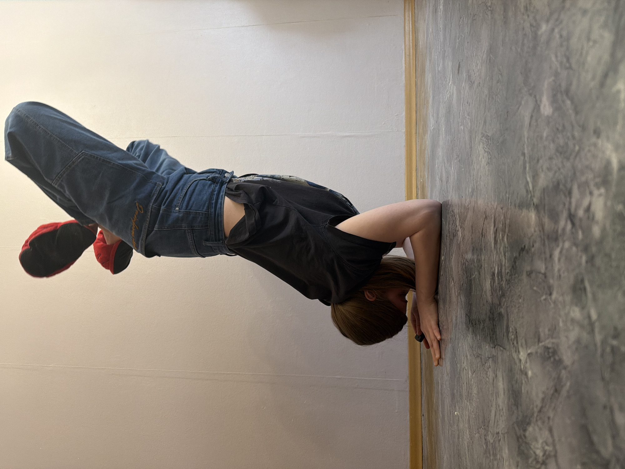 The image shows a person performing a headstand or forearm stand indoors. The individual, with light brown hair, is balanced on their head and forearms on a dark grey and black textured floor. Their body is inverted, with legs extended upwards, leaning against a plain, light-colored wall.
The person is dressed in dark blue baggy jeans and a black t-shirt. Visible on the back right pocket of the jeans is light-colored embroidery that spells "Doodyz." They are wearing red, slipper-like shoes with black soles. A thin, light brown wooden trim or door frame runs vertically where the floor and wall meet on the right side of the frame. The setting appears to be a simple room with functional lighting. The person's face is obscured as it is pressed against the floor. The original photograph was taken with a rotated camera, presenting the inverted pose horizontally.