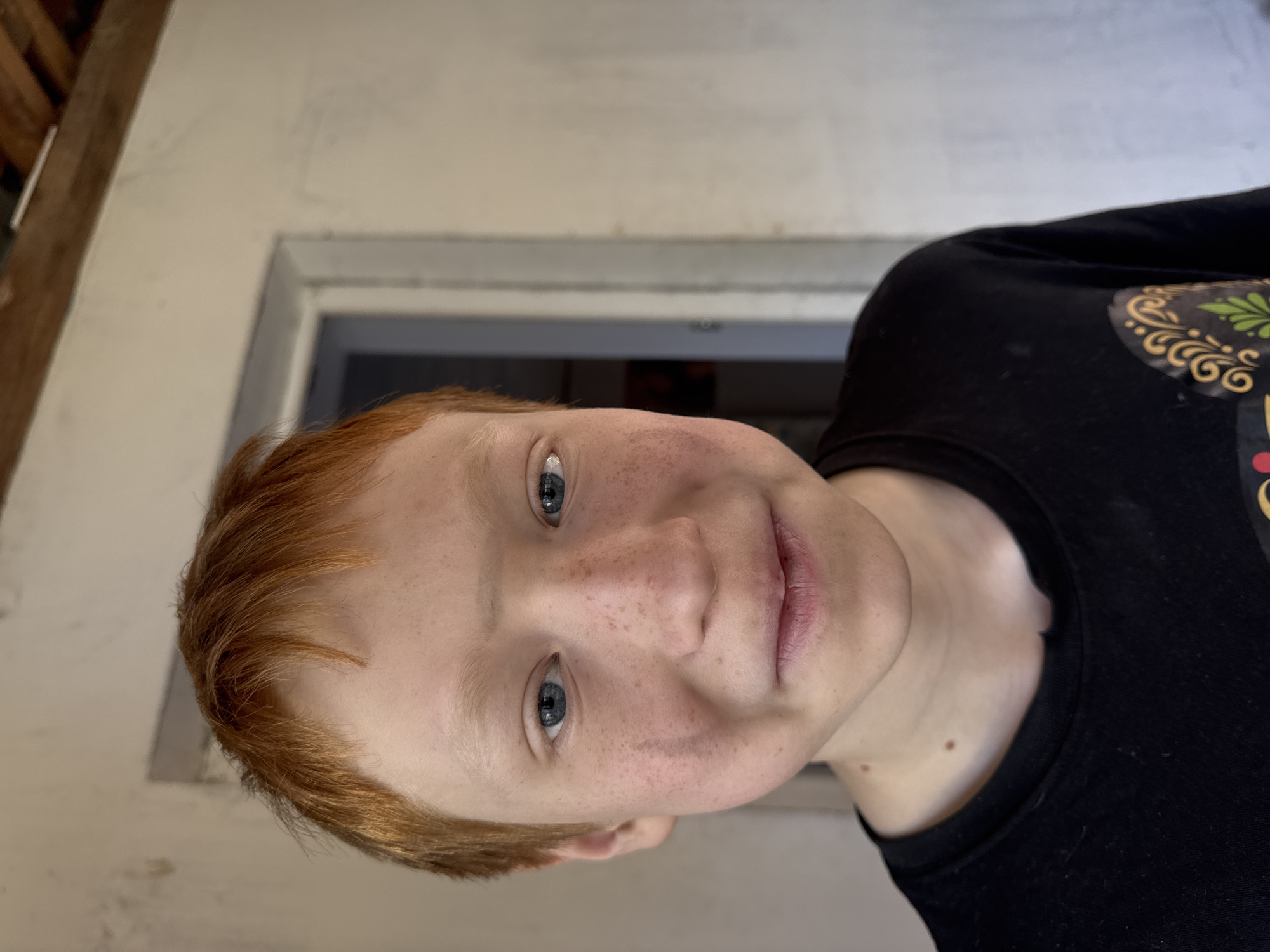 A young boy with red hair and blue eyes is seen in a close-up, slightly rotated view. He has fair skin with freckles and is wearing a black T-shirt with a colorful design on the shoulder. The background is blurry and appears to be an interior setting with a white wall and a doorway. There is no information to confirm that this image was taken in the city of Chaikovsky, Russia.