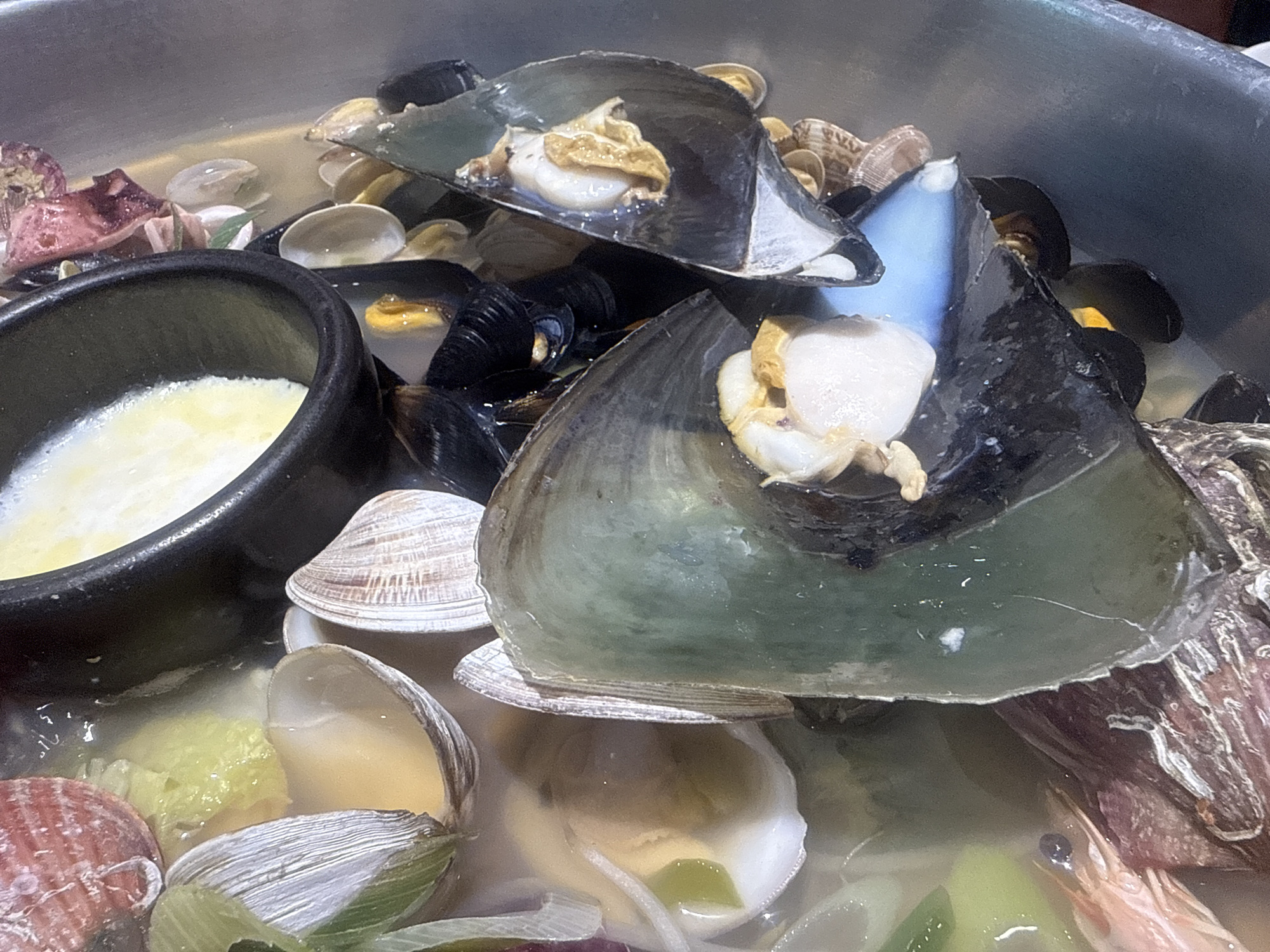 The image shows a stainless steel pot filled with a seafood stew. The stew contains a variety of shellfish, including open clams with visible meat, mussels, and large, open shellfish with a white, fleshy interior and dark green shells. A small black bowl filled with a pale, creamy substance sits near the left edge of the pot. A partially visible shrimp rests on the right side of the frame. The overall setting suggests a Korean seafood dish. The location is Gimpo-si, South Korea.