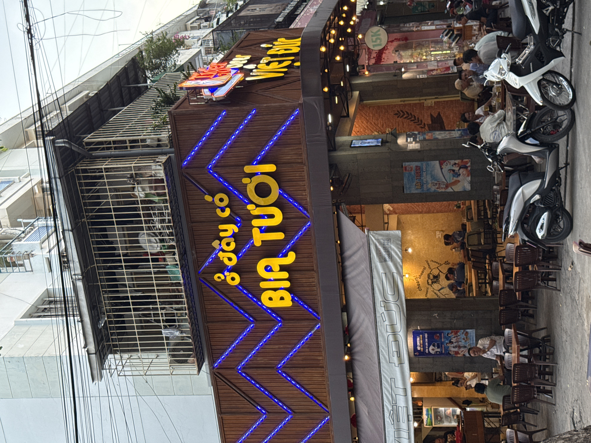 This image depicts a street-level view of a restaurant or bar in Nha Trang, Vietnam, with a prominent sign reading "BIA TUOI" (fresh beer) illuminated by blue LED lights. The establishment has an outdoor seating area with numerous tables and chairs, and several people are seated, engaged in conversation or eating. Motorcycles are parked in front of the establishment. The building features a second-story balcony with a metal cage. A banner above the outdoor seating area displays the text "VIET DUC" in large letters. The overall scene suggests a lively urban environment characteristic of Vietnam.