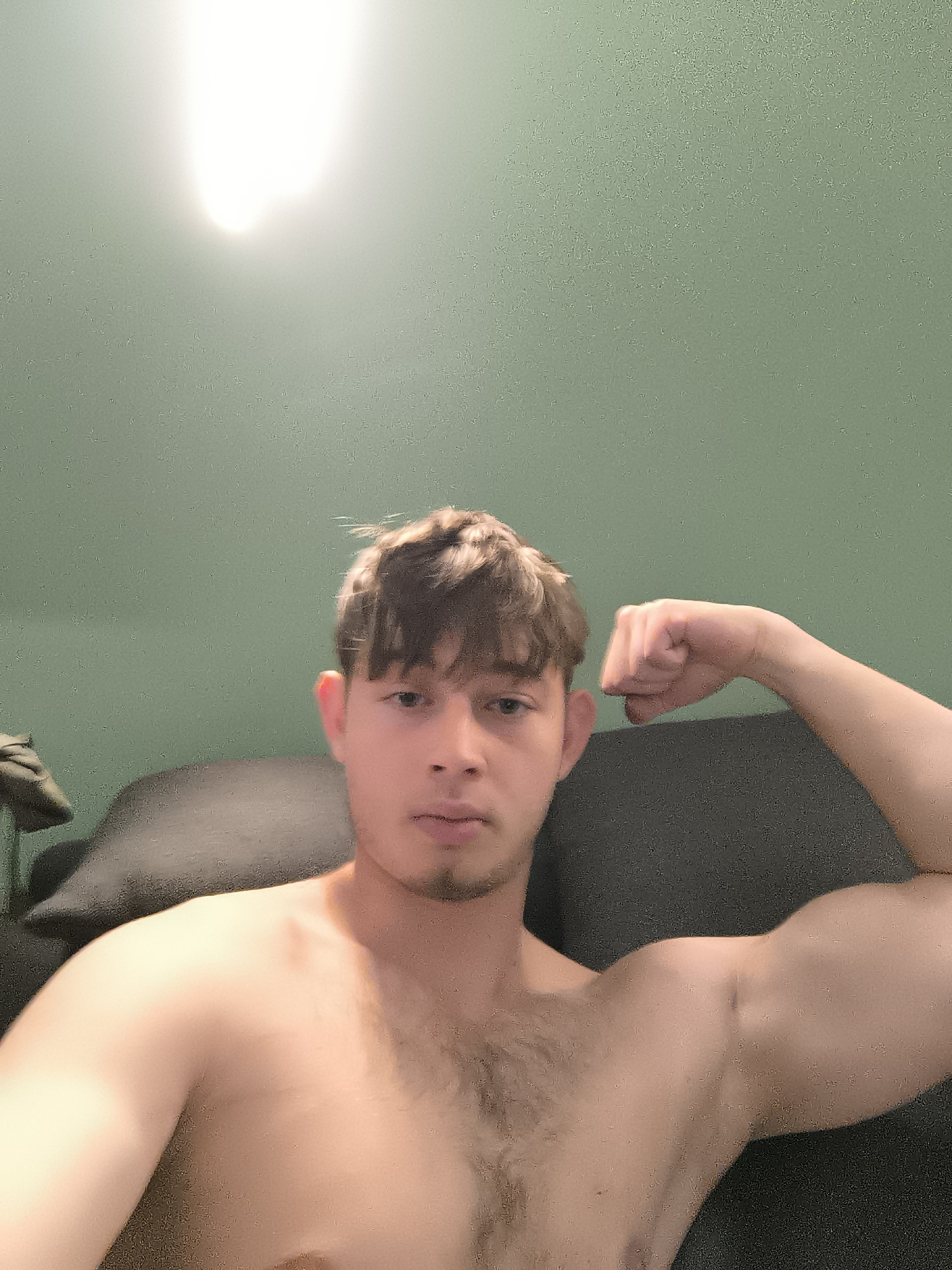 A shirtless young man is flexing his right bicep while seated on a dark grey couch. The background is a light green wall with a bright light source positioned above the subject. The photograph appears to be taken indoors. There is no evidence to suggest that the image was taken in the city of Sofia, Bulgaria.