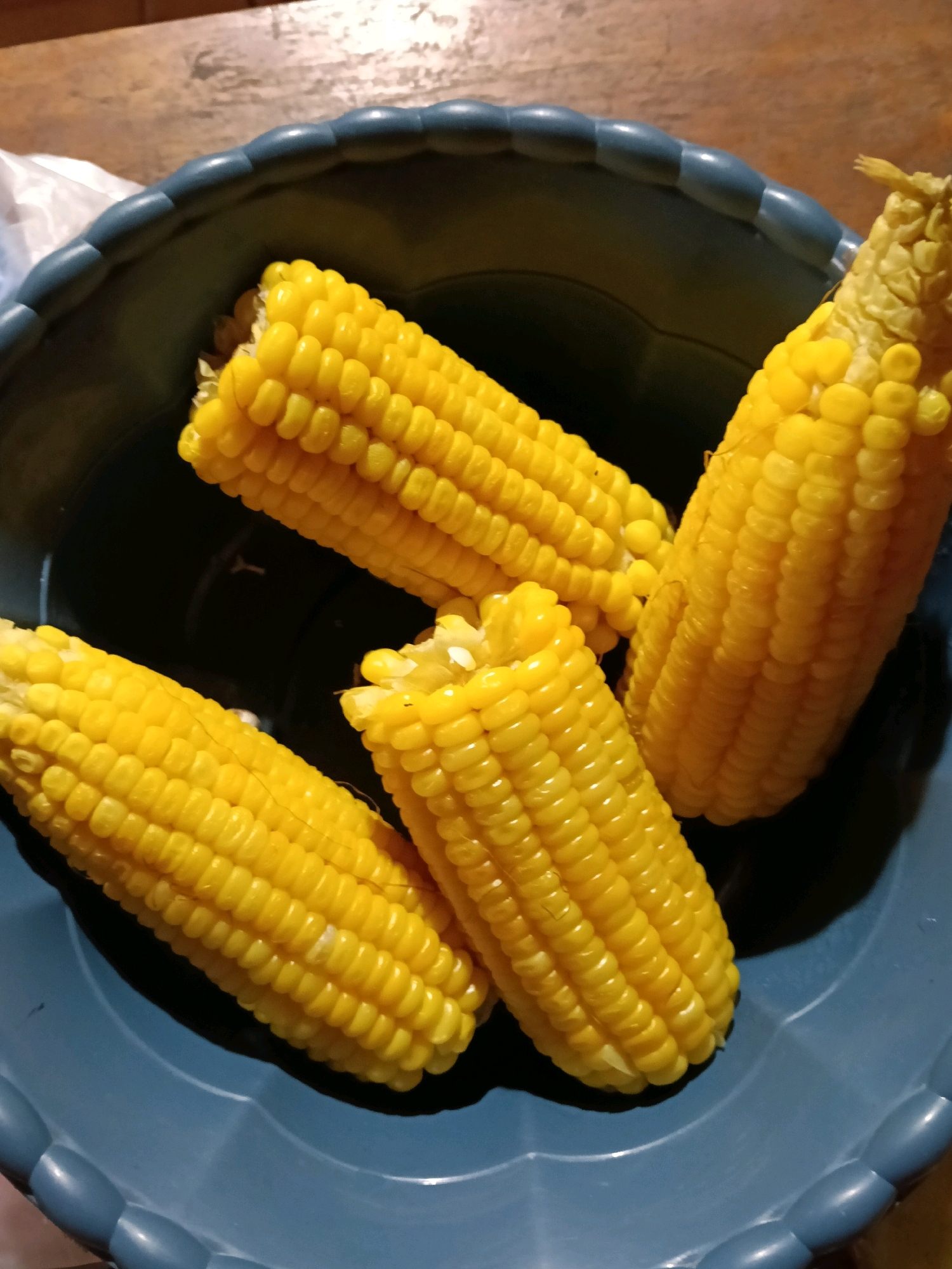 Four ears of cooked corn are arranged in a dark blue plastic bowl. The corn is bright yellow and appears to have been recently cooked, with some kernels showing slight separation. The bowl is textured with a scalloped edge and sits on a wooden surface. A white plastic bag is visible to the left of the bowl, partially obscured. The scene is lit from above, casting shadows within the bowl. The image was taken in the city of Galuk, Indonesia.