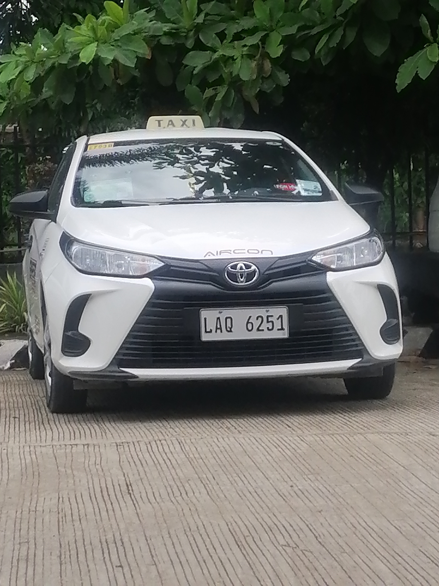 A white taxi is the central subject of this full-frame, eye-level shot, parked outdoors on a patterned concrete surface. The car is a Toyota sedan, identifiable by its emblem on the grille. A "TAXI" sign is affixed to its roof, and "AIRCON" is visibly printed on the car's hood, above the grille. The vehicle's license plate, located on the front bumper, displays the alphanumeric combination "LAQ 6251".
The car is clean and appears to be in good condition. Various stickers are visible on the windshield, though their details are mostly indistinct, save for a yellow sticker with faint text "E700X" on the driver's side. Large, lush green foliage forms a dense backdrop behind the vehicle, suggesting a tree-lined street or a garden setting. The lighting is soft and diffused, indicating either an overcast day or the car being parked in a shaded area, possibly early morning or late afternoon, as there are no harsh shadows. No people are visible in the image, and the taxi appears to be stationary. The overall scene suggests a typical daytime setting in an urban or suburban area, consistent with the provided location context of Davao City, Philippines.