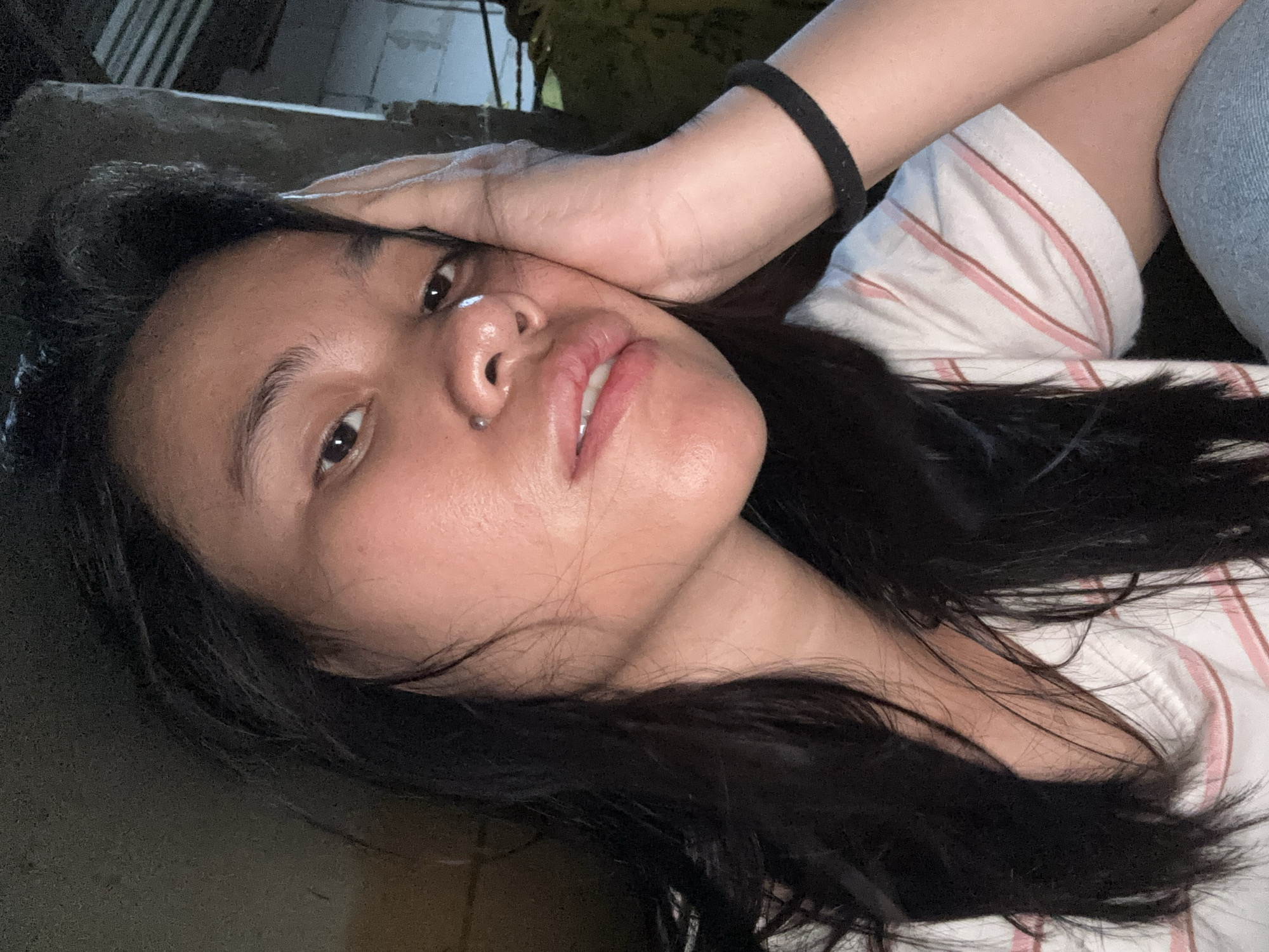 In a close-up, low-light selfie taken in East Jakarta, Indonesia, a person with dark, shoulder-length hair rests their right hand against their face, with their palm supporting their cheek. They are wearing a white t-shirt with thin pink and red stripes. A black wristband is visible on their right forearm. The background is dark and out of focus, with hints of a textured wall and possibly some corrugated metal structure. A small silver stud is visible on the person's nose. The lighting is dim, creating a moody atmosphere.
