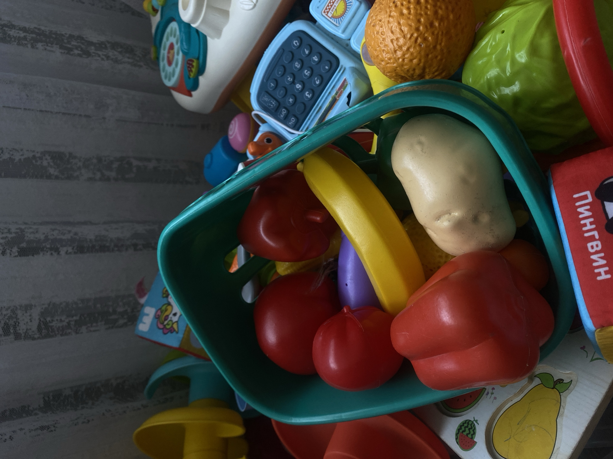 In an indoor setting, likely a child's play area, a collection of plastic toy food items is piled together. The dominant colors are bright and varied, with a prominent teal basket holding several red toy vegetables, including what appear to be tomatoes and a bell pepper. A yellow banana-shaped toy is also visible, along with a light-colored toy potato. Other toys scattered around include a blue toy calculator, a toy orange, and a green toy cabbage. The background features a textured, light-colored curtain with vertical stripes and a hint of a patterned tablecloth or playmat with fruit designs. The scene is well-lit, with shadows suggesting a light source from the right side of the frame. The toys appear to be for a young child's play and are not associated with any specific activity or interaction. The location of Пешнево, Russia is not determinable from the visual information present in the image.