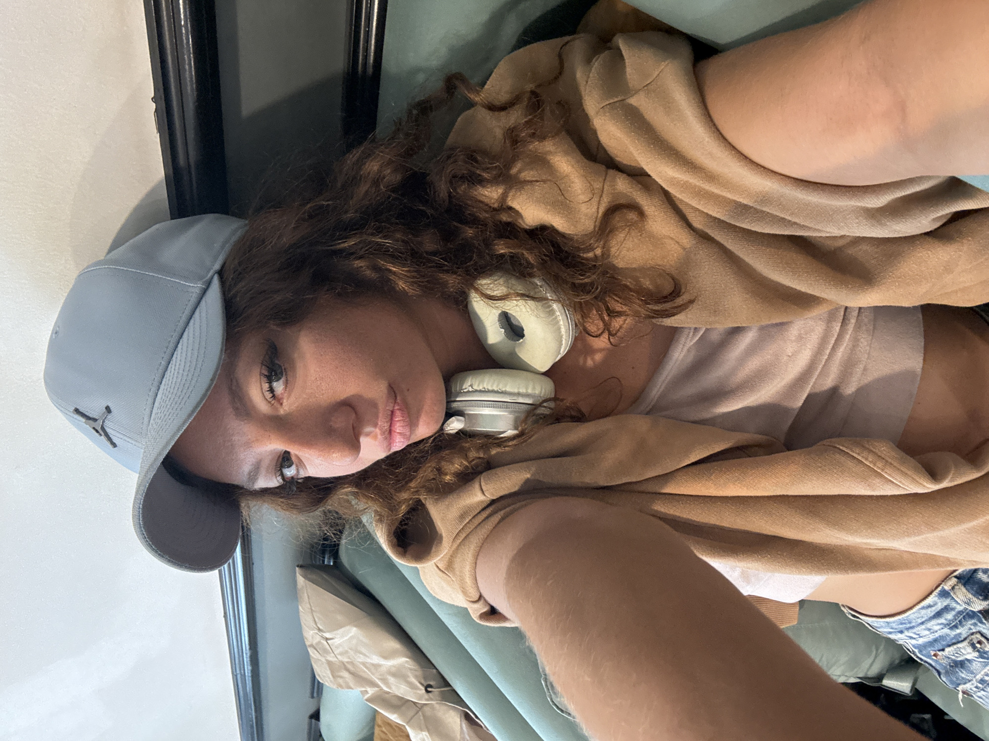 A woman with curly brown hair is wearing a light blue baseball cap and beige headphones around her neck. She is wearing a tan-colored hoodie and a light pink top. Part of a denim garment is visible. The setting appears to be indoors.