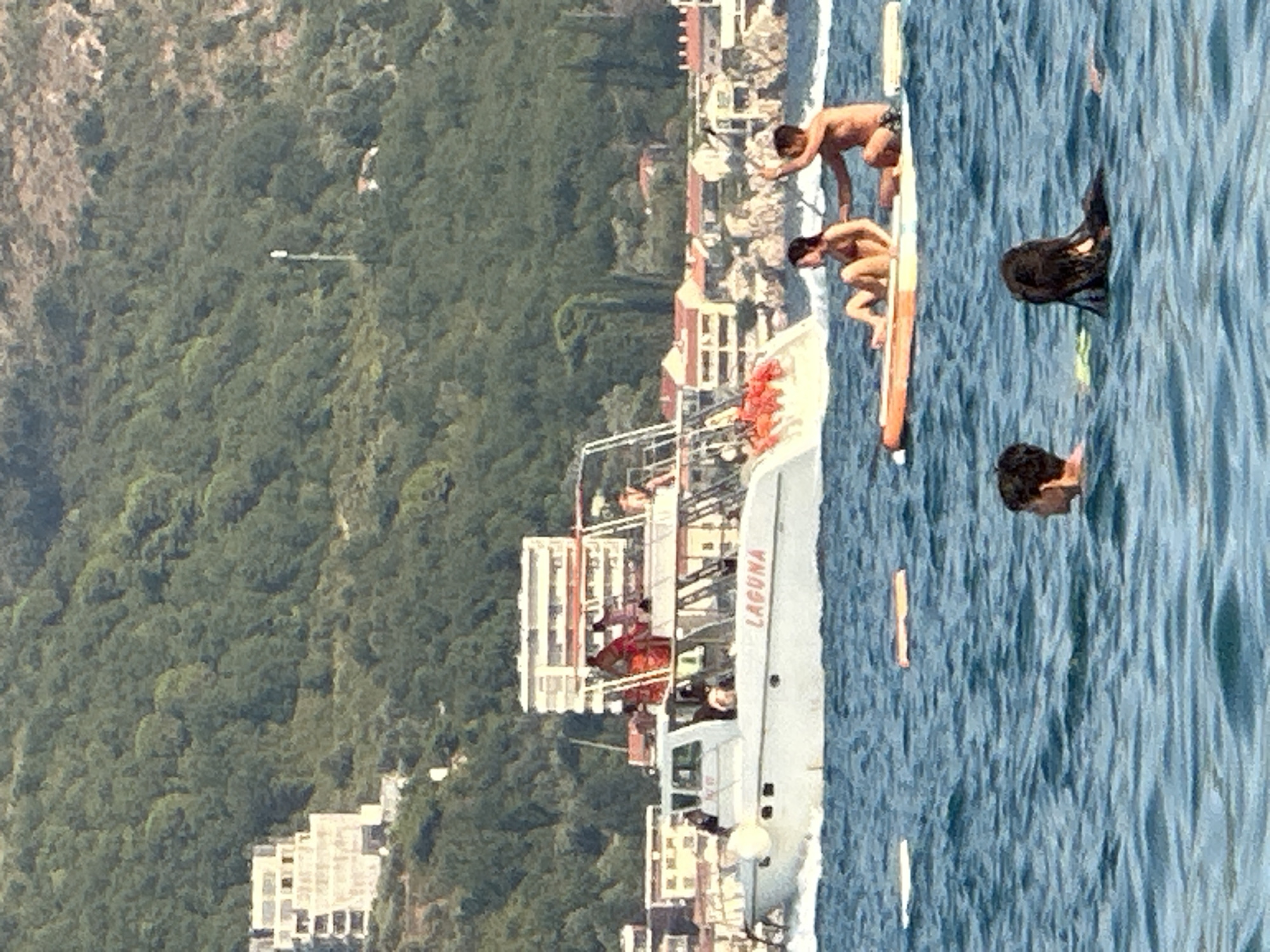 In the foreground, two individuals are partially submerged in the choppy blue water. To the left of them, another person's head is visible above the water. In the mid-ground, a white boat with the name "LAGUNA" in red lettering is cruising on the water. Several people are on the boat, some wearing orange life vests. The boat has a white cabin with windows and a larger open deck area towards the stern. Further in the mid-ground, two people are sitting on a paddleboard, with one person appearing to be paddling. In the background, a coastline is lined with buildings, some appearing to be hotels or apartments, built on a hill covered in lush green trees. The hill slopes upwards towards the top of the image. The sky is a bright, clear blue. The overall scene depicts a sunny day with recreational activities taking place on the water near a coastal town.