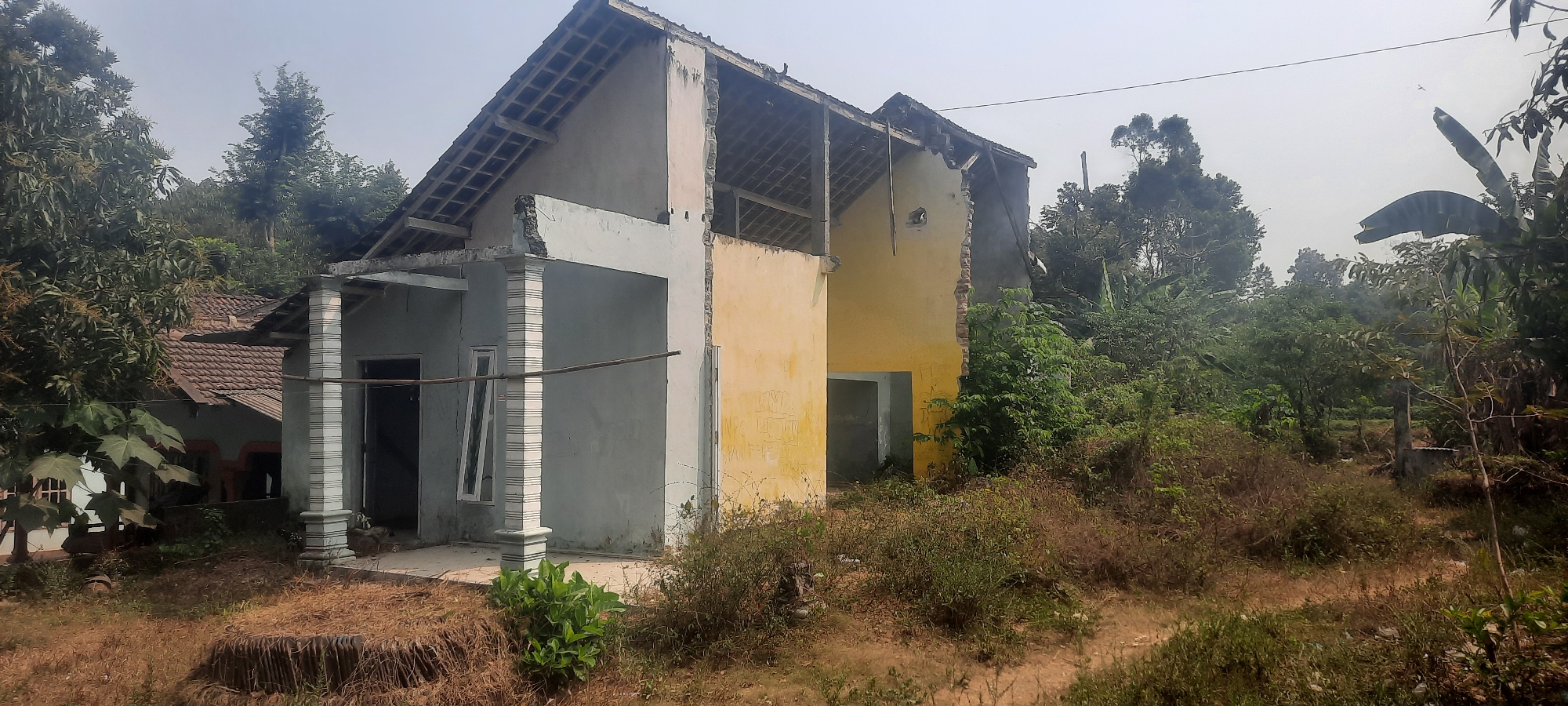 The image shows a partially demolished house with its roof structure exposed. The house appears to be in a state of disrepair, with one side of the yellow-painted wall significantly broken. The area surrounding the house is overgrown with vegetation, including dry grass, bushes, and trees. In the background, more trees and foliage can be seen, suggesting a rural or semi-rural environment. A smaller house with a tiled roof is partially visible to the left of the damaged structure. The sky is overcast and appears hazy. The scene is located in Ploso, Indonesia. There are no people visible in the image.