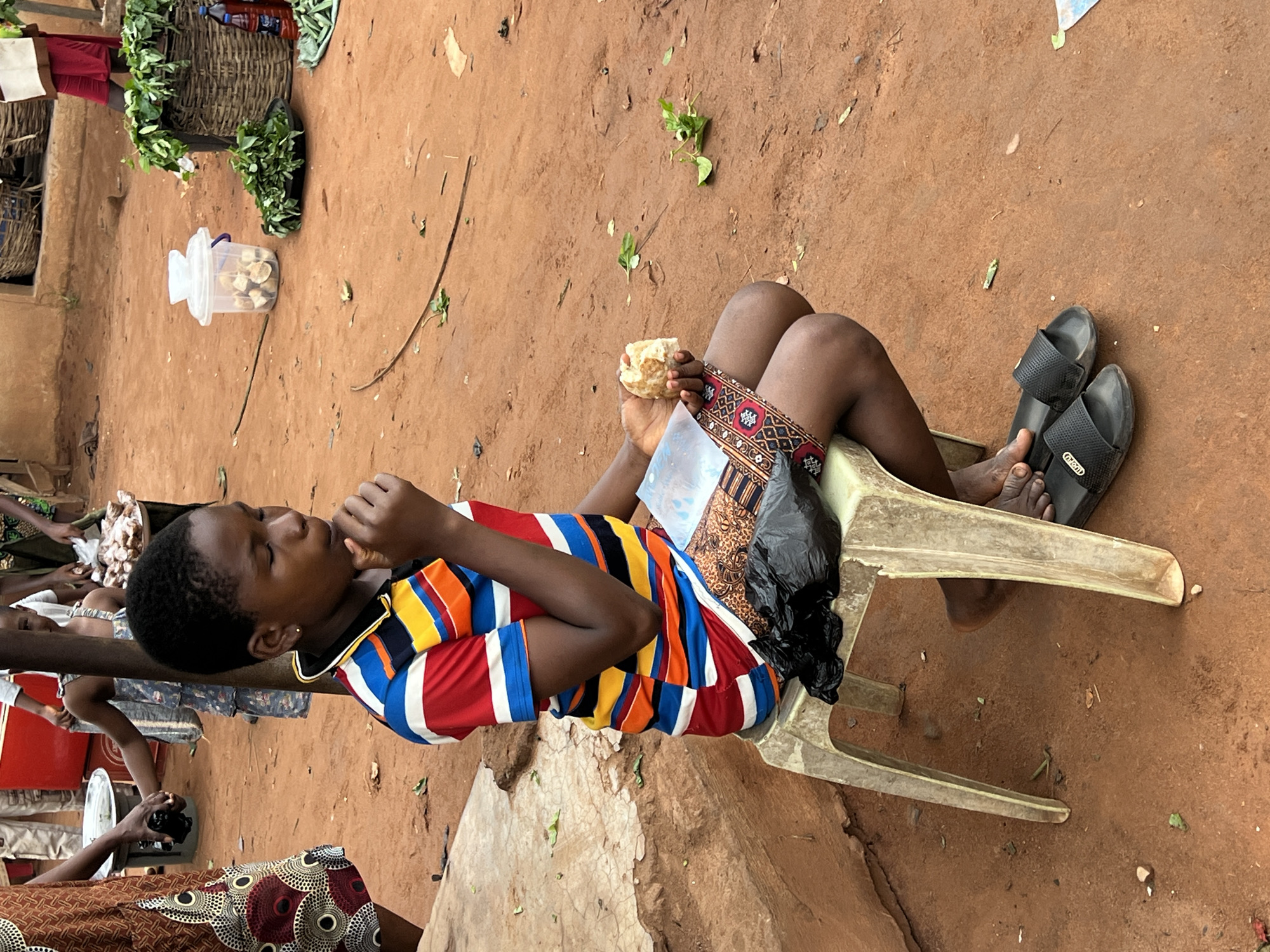 In Nkpor, Nigeria, a young Black individual sits on a beige plastic stool, eating a piece of bread. They are wearing a horizontally striped shirt in red, blue, yellow, and white, and a patterned skirt. The ground is made of red earth, with scattered leaves and debris. In the background, several individuals are visible, some tending to market stalls. Objects like woven baskets, a plastic container with food items, and bottles are present. The scene appears to be an outdoor market setting.