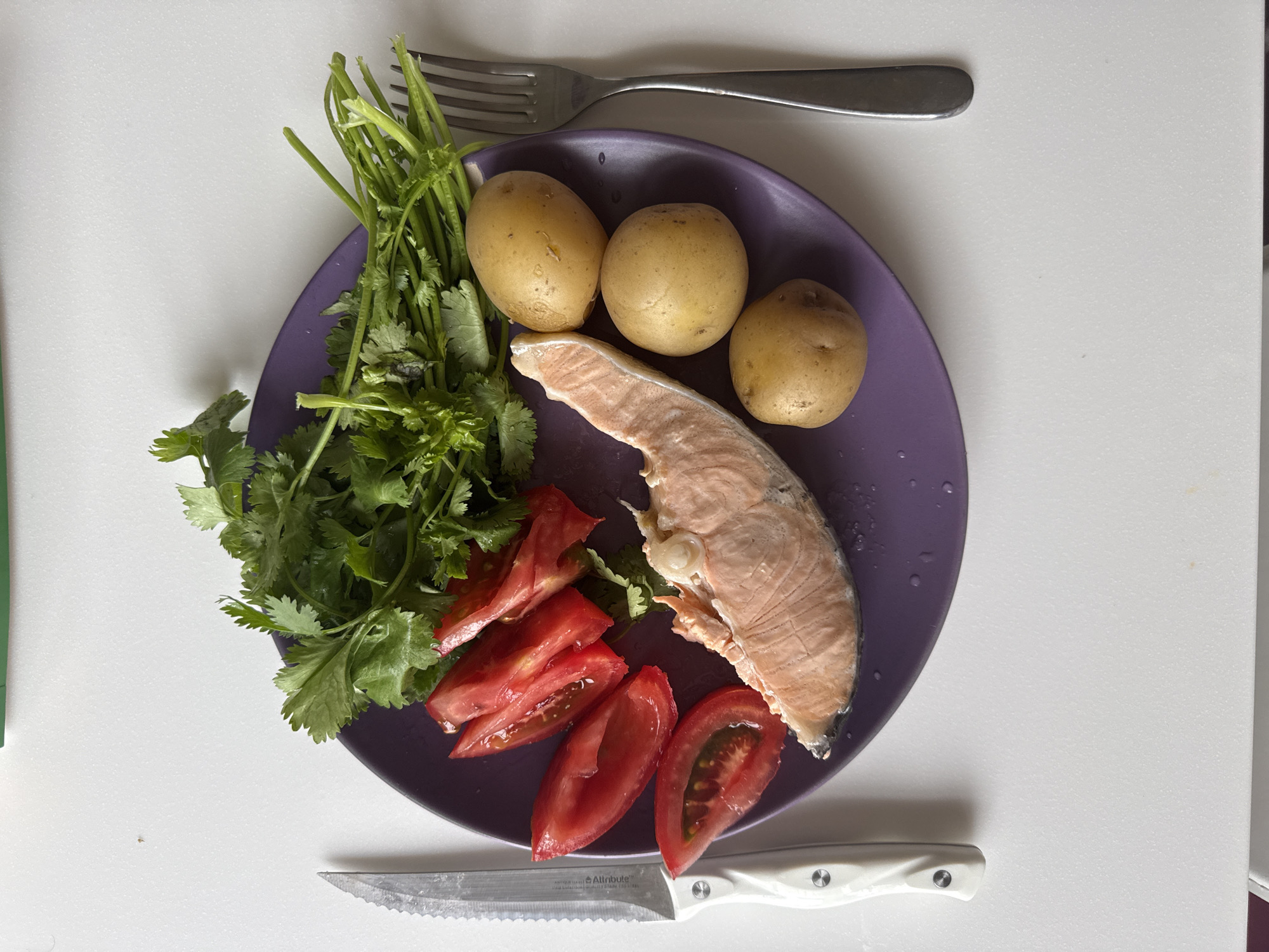 On a white surface, a purple plate holds a cooked salmon fillet, sliced tomatoes, three small potatoes, and a bunch of cilantro. A serrated knife with a white handle rests above the plate, and a fork lies below it. The scene is lit from above, casting minimal shadows. There are no people or animals present. The background is a plain white surface, with a faint greenish object partially visible on the right edge. There is no indication of the specific location within Zimyonki (Zimenki, Zimenka), Russia.