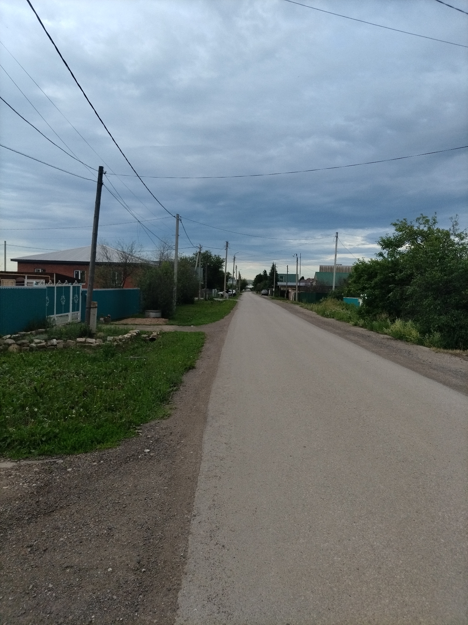 The image depicts a rural road in Nizhnyaya Makatama, Russia, on an overcast day. A paved road stretches into the distance, flanked by grassy verges and gravel shoulders. On the left side of the road, a turquoise metal fence encloses a property with a brick house with a silver roof. Several utility poles with overhead wires are visible along the roadside, indicating electricity or communication infrastructure. Trees and bushes line the properties and the opposite side of the road. A few cars are parked or driving in the distance, suggesting a residential area. The sky is filled with heavy, gray clouds.