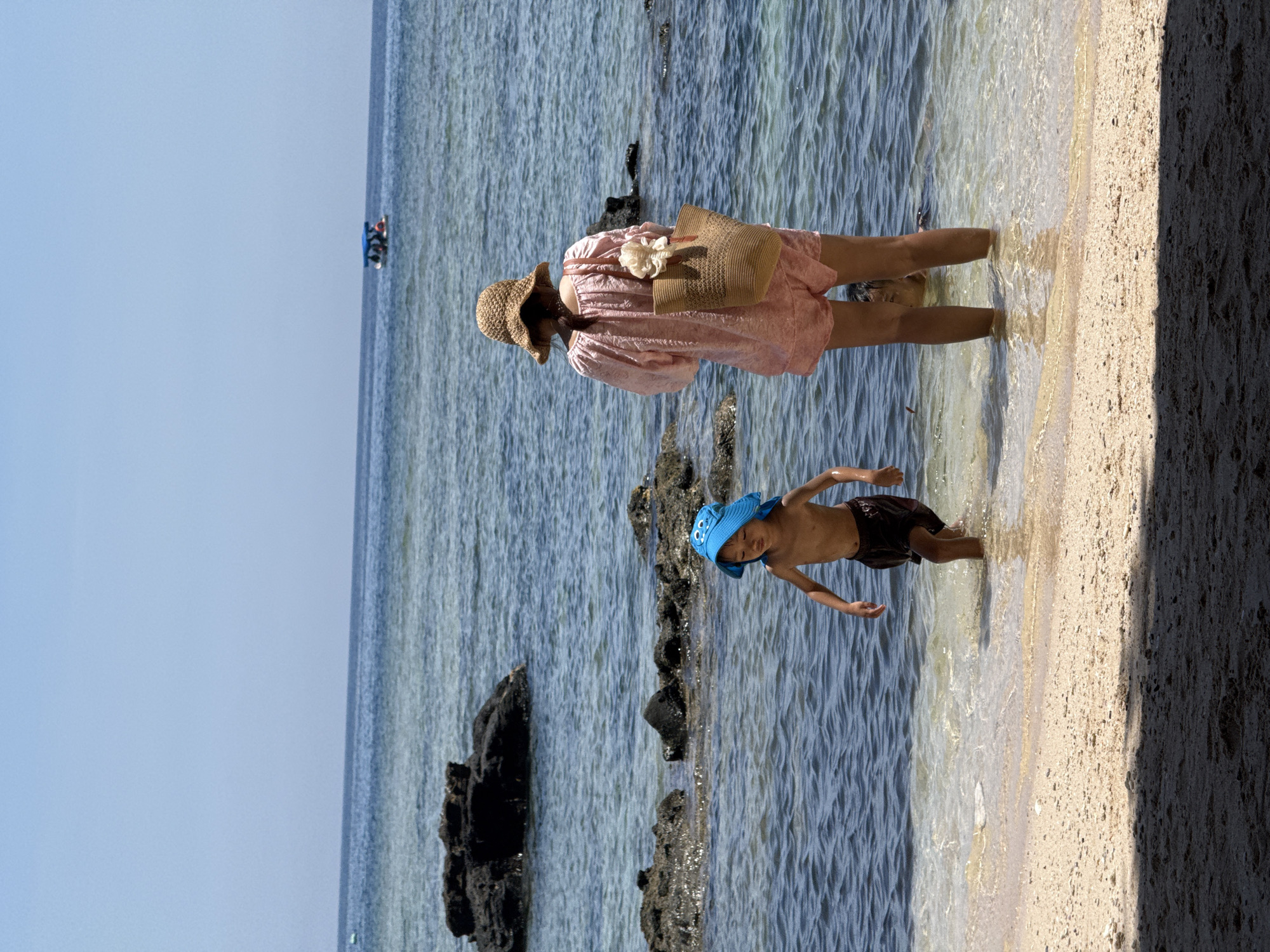 Two people, an adult and a child, are standing at the edge of the water on a sandy beach, with the sea and a boat in the background. The adult, viewed from the back, wears a straw hat and a pink outfit, carrying a straw bag. The child, facing left, is wearing a blue sun hat and dark swim trunks, with their upper body bare. They are in shallow, clear water. The environment is a beach with dark rocks visible in the water and a bright blue sky. The scene appears to be on Lý Sơn Island, Vietnam, a location known for its beaches and clear waters.