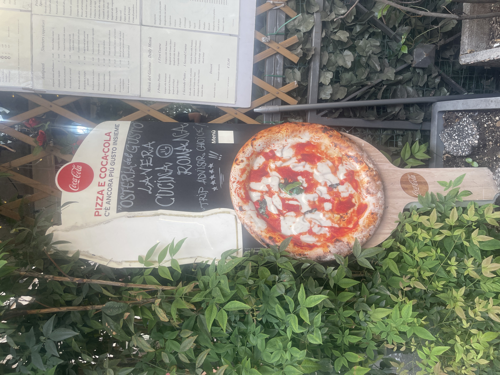The image shows a sign outside a restaurant in Rome, Italy. The sign is painted with the Coca-Cola logo and text in Italian that includes "PIZZA E COCA-COLA," "OSTERIA del GUSTO," "LAVERA," and "CUCINA ROMANA." The sign also features a five-star rating and "TRIP ADVISOR CHOICE." To the right of the sign is a large image of a pizza on a wooden platter with the Coca-Cola logo. The sign and pizza are partially obscured by green foliage. In the background, there is a menu board listing antipasti, first courses, second courses, and desserts. A wooden lattice fence is behind the menu. A flower pot is visible to the right.