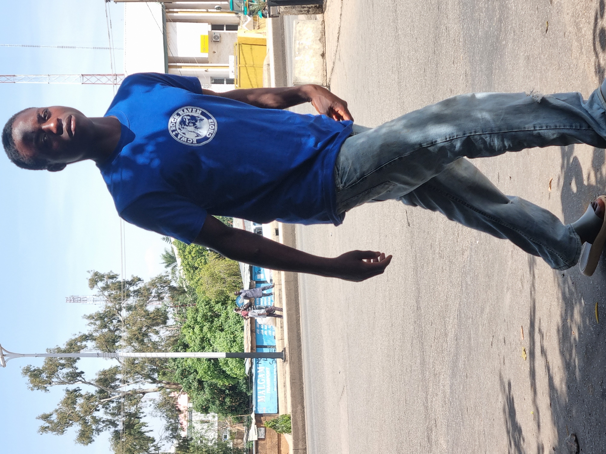 A young Black man stands on a street in Jos, Nigeria, facing left and angled slightly toward the camera. He wears a blue t-shirt with a circular logo and light-colored jeans. The man is positioned on a grey road near a sidewalk, with buildings and trees visible in the background. Other people can be seen standing near a sign in the background. A tall, metal telecommunications tower is visible in the background as well. The weather appears sunny and bright.