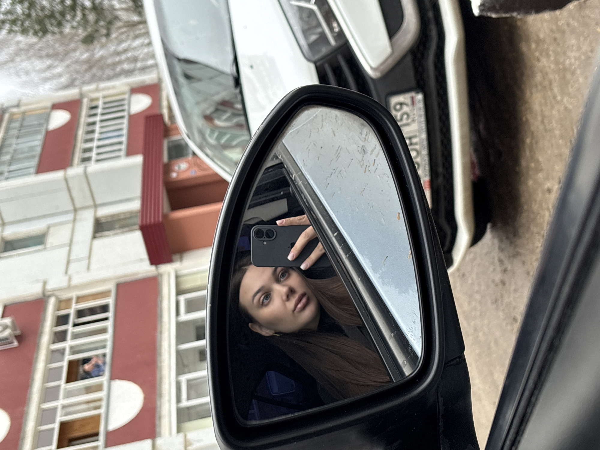 A woman is taking a selfie in the side mirror of a car. The car, a white hatchback, is parked next to a building in Chaikovsky, Russia. The building has a red and white facade with multiple windows, some with visible people inside. The license plate of the vehicle in front of her is visible.