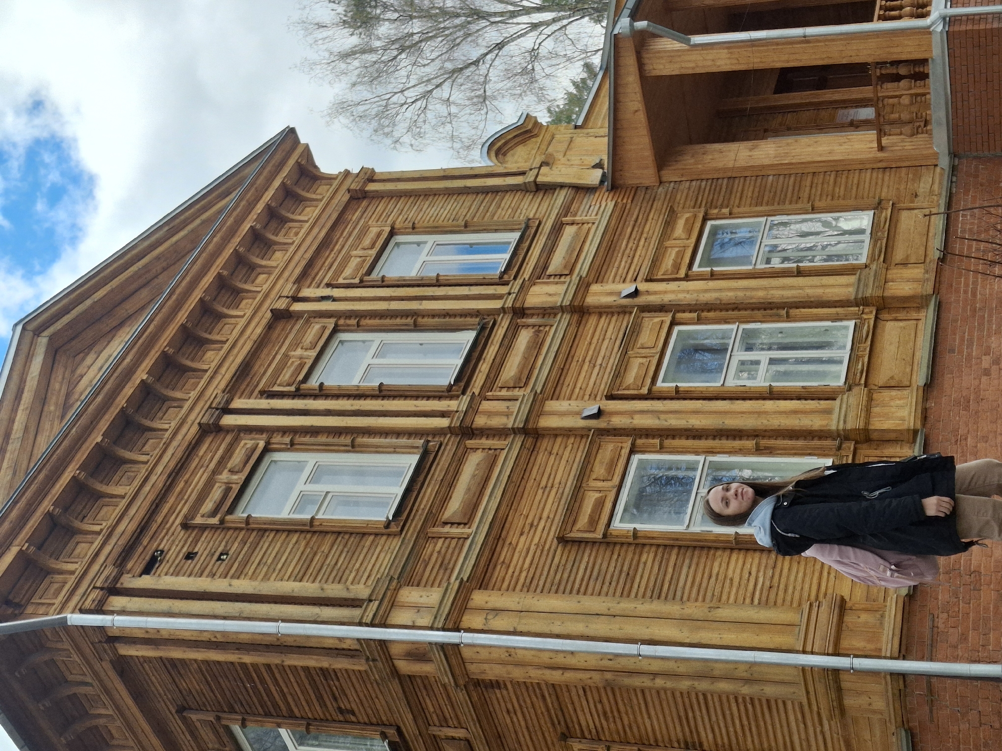 A wooden building with detailed trim work stands prominently, featuring multiple windows on each level. A young woman in a black jacket and beige pants stands in front of the building on a brick section. The sky is visible, with blue patches and clouds, and there is a bare tree branch above. The scene suggests a location in Sarapul, Russia, judging by the architectural style of the building.