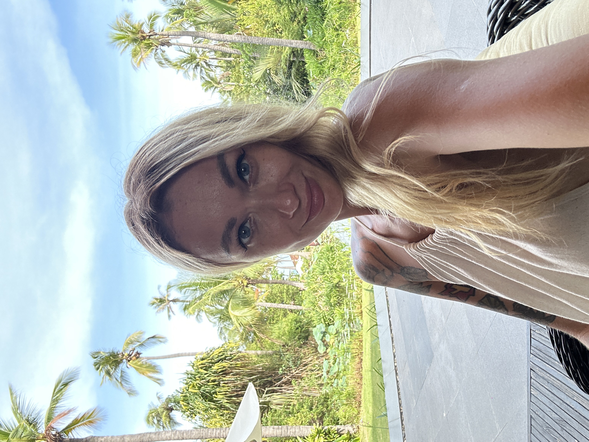 A selfie of a blonde woman with blue eyes and fair skin is taken outdoors. She is wearing a light-colored, strappy dress. Her left arm and shoulder are visible, showing a tattoo. She is seated in a woven chair. Behind her, lush green foliage and palm trees are visible under a bright blue sky with some clouds. The foreground shows a tiled patio and wooden decking.
