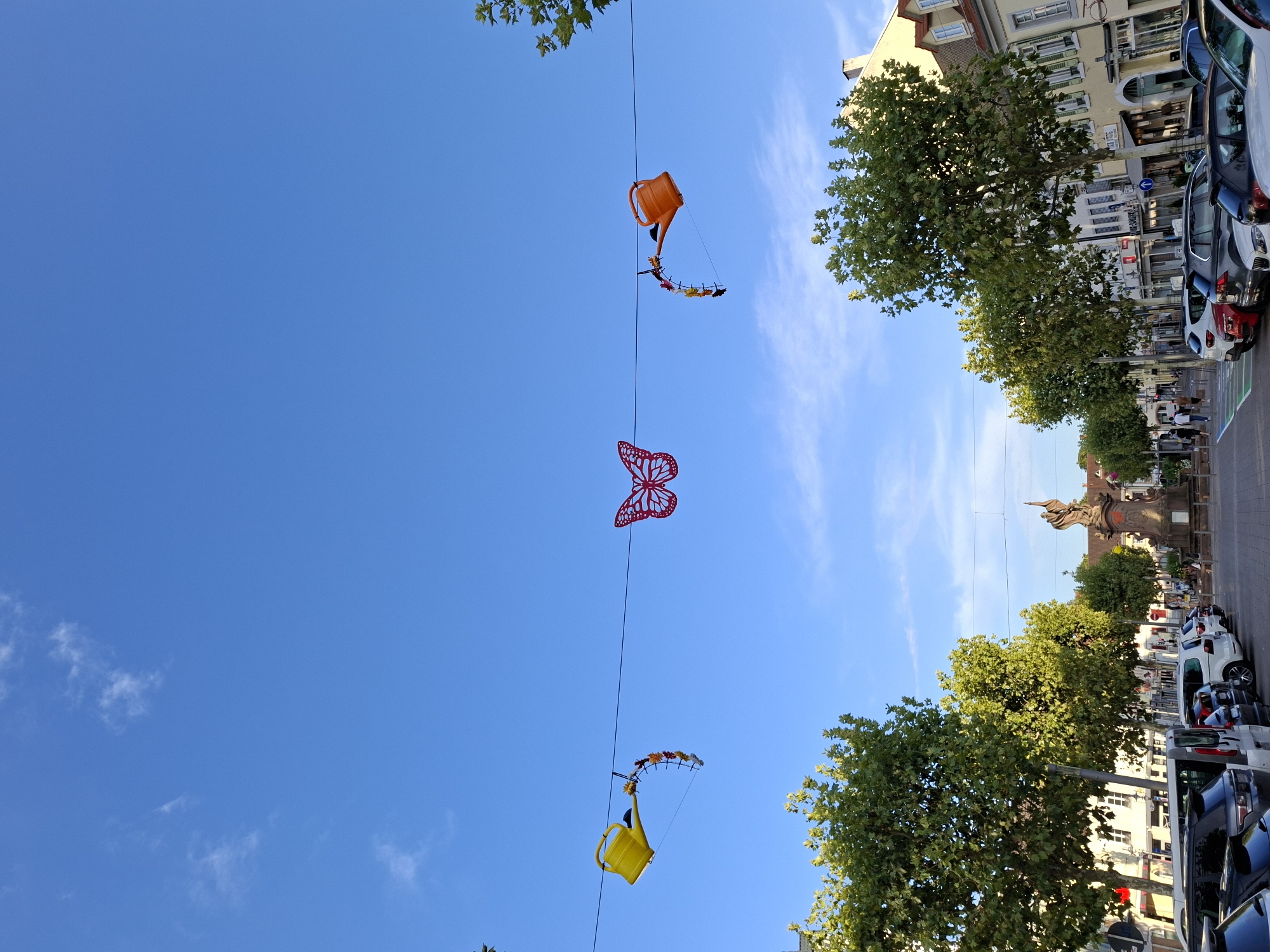 The image shows a street scene in Rastatt, Germany. The sky is clear blue with a few wispy clouds. Overhead, a wire is strung across the street, decorated with a large red butterfly in the center and two watering cans on either side. The watering cans appear to be part of a larger decoration, with a circular frame adorned with what look like small artificial flowers or leaves hanging from them.
Below the decorations, a row of leafy green trees lines the street. Beyond the trees, a street with parked cars is visible, leading towards a plaza with a statue in the background. Buildings with traditional architecture, featuring light-colored facades and dormer windows, line the street on the right side of the image. The street appears to be a public space, possibly a pedestrian zone or a street with limited vehicle access, as indicated by the presence of a statue and what might be a fountain base at the far end of the plaza. The overall impression is of a pleasant, sunny day in a European town square.
