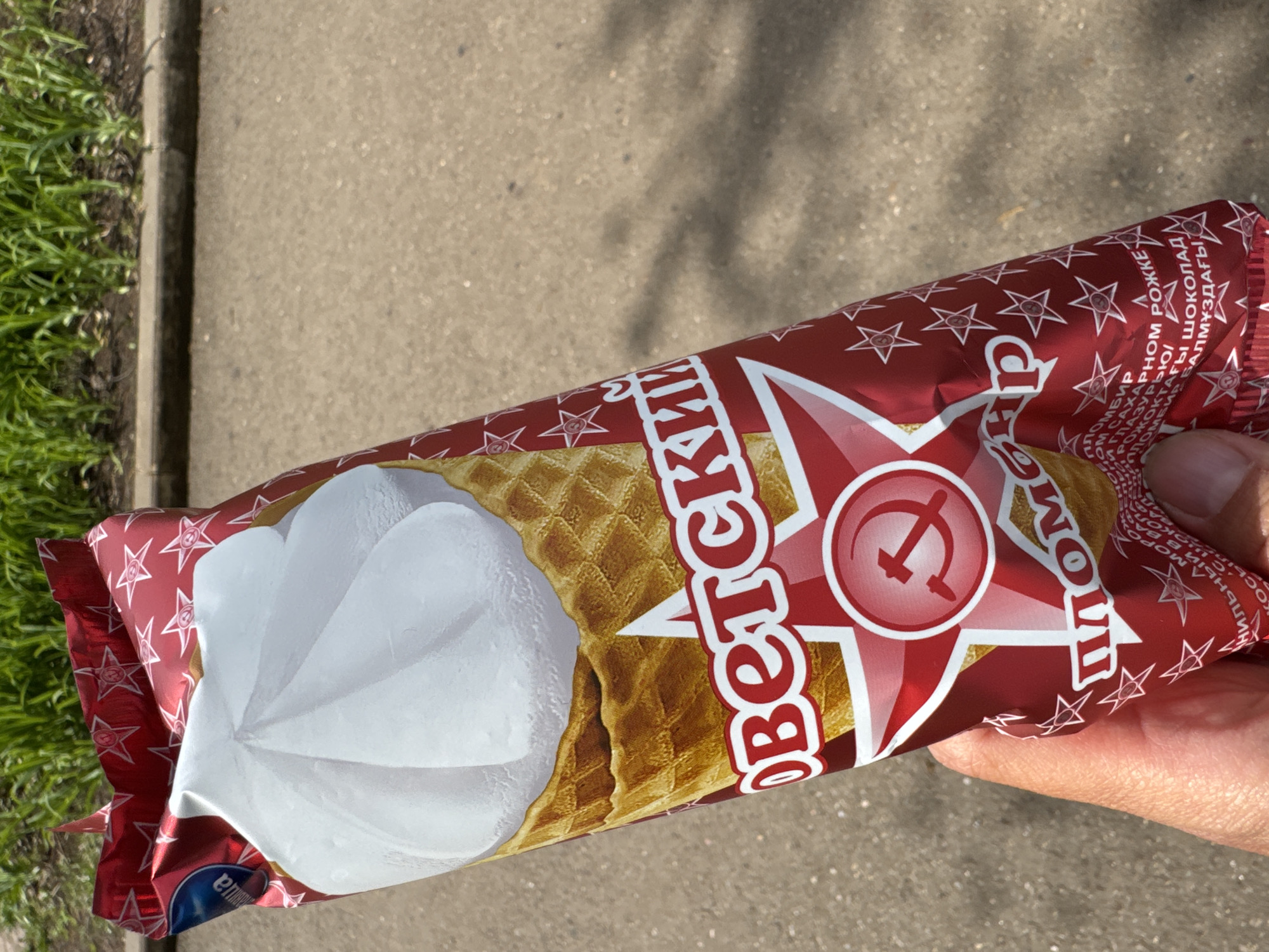 A person is holding a Soviet-branded ice cream cone in its packaging, which features a red and white design with stars and a stylized image of the ice cream. The packaging is labeled with text in Russian, including "Soviet" and "ice cream." The setting appears to be outdoors, with pavement and grass visible in the background. The image was taken in the city of Chaikovsky, Russia.