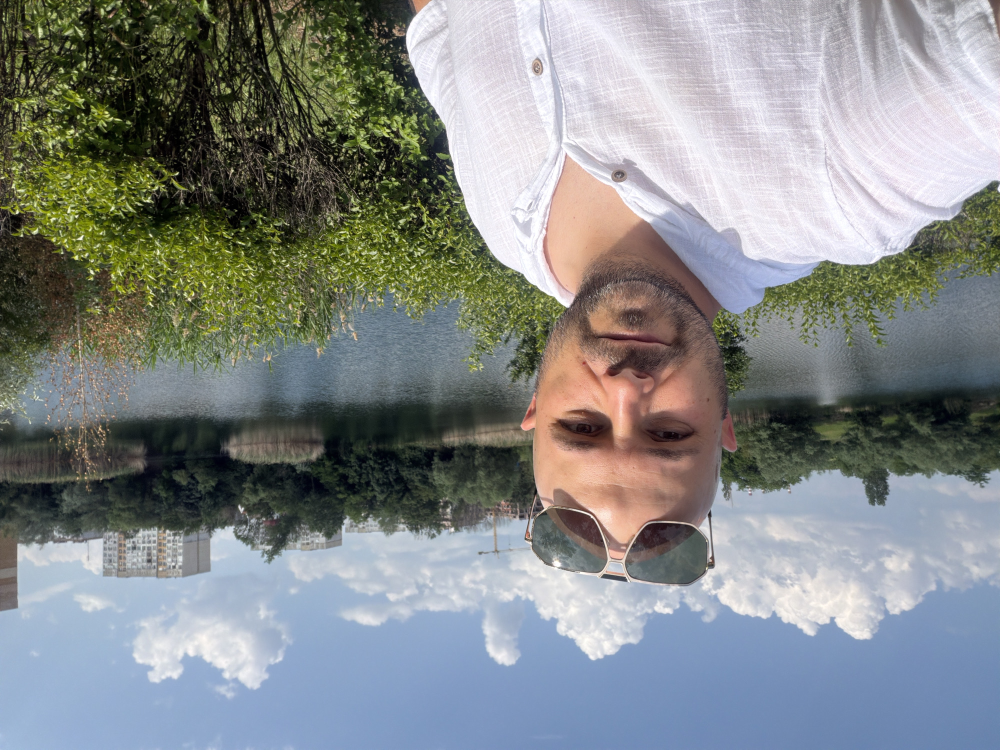 A bald male individual, with a light-colored beard, is depicted in a close-up outdoor selfie. He wears dark sunglasses perched on his forehead and a white, textured, short-sleeved linen shirt with wooden buttons. His gaze is directed towards the camera.
In the mid-ground, behind the individual, a calm body of water, likely a lake or large pond, extends across the frame, reflecting the bright blue sky. The water's edge is lined with green foliage, including dense bushes in the foreground on the right, and reeds and trees on the far bank.
The background reveals an urban landscape beyond the distant treeline. Several multi-story residential apartment buildings, predominantly light grey with some yellow accents, are visible. To the left of these buildings, a tall yellow construction crane rises above the trees, indicating ongoing development. The sky above is clear and blue, dotted with scattered white cumulus clouds, suggesting a sunny day.
The architectural style of the distant buildings suggests a location consistent with urban areas in Eastern Europe, but a precise city cannot be determined from the visual evidence provided. Therefore, the location in the city of Unknown, Unknown cannot be specifically identified.