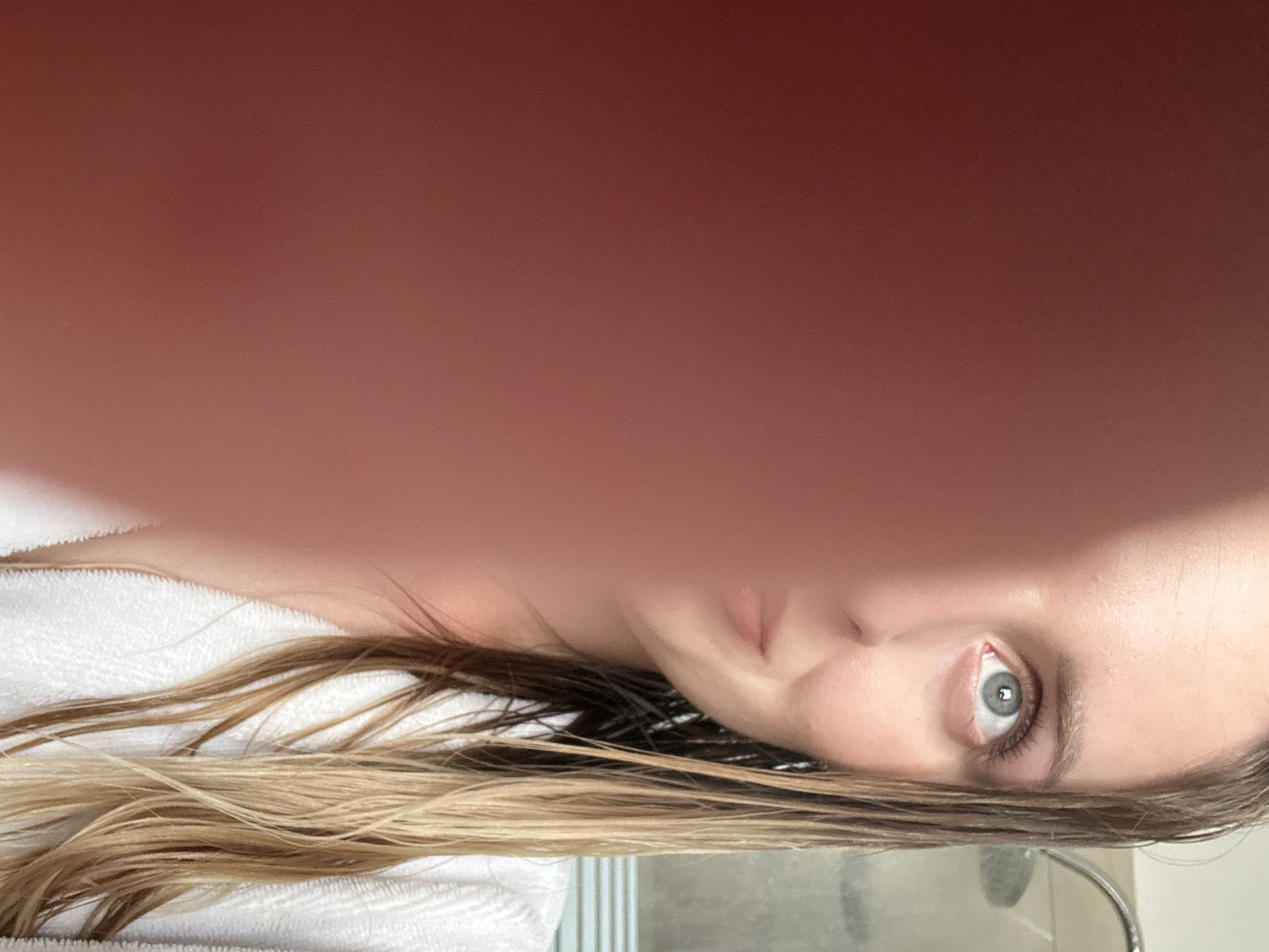 A young woman, partially obscured by a reddish-pink blur that appears to be a hand or arm, is looking upwards and slightly to her right. Her light-colored, wet hair is visible, framing her face. She is wearing a white bathrobe. In the background, out of focus, is a shower head, suggesting the setting is a bathroom. The image is a selfie. The location in Petah Tikva, Israel, is not discernible from the visual information provided.
