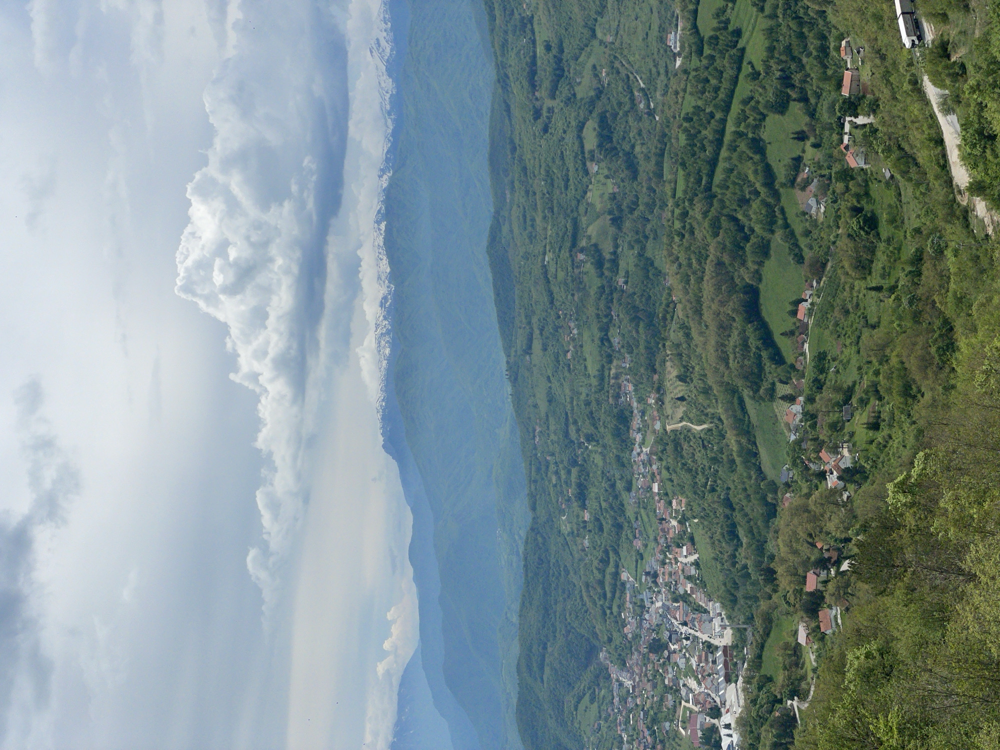 The image shows a landscape in Gmići, Bosnia and Herzegovina. On the left, there are clouds in the sky. Next to the clouds is a mountain range. A dense, green forest covers the rest of the landscape, with a town nestled between the trees and hills. At the top right, a road winds through the forest with at least one car on it. Scattered buildings are visible throughout the wooded area.