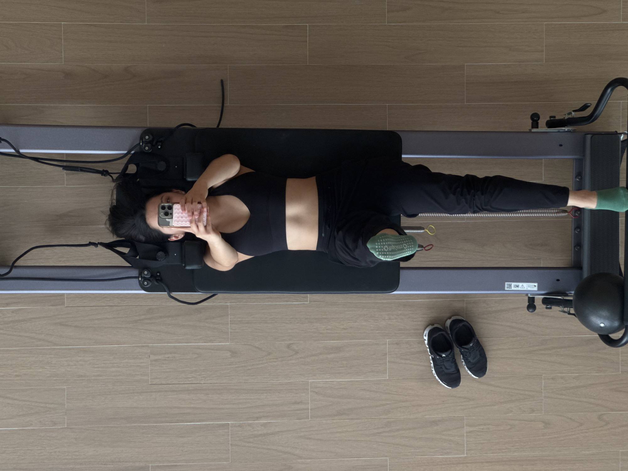 A bird's-eye view shows a woman lying on a Pilates reformer machine indoors. The woman has dark hair and is wearing a black sports bra and black pants. She is holding a phone with a pink case in front of her face. She has green socks on her feet. The reformer machine is gray with a black carriage. A pair of black sneakers are on the floor next to the machine. The floor is covered with light brown wood-like tiles. The location is unknown, but the prompt suggests it may be in San Bernardino Tlaxcalancingo, Mexico. The scene appears to be taking place in a gym or fitness studio.
