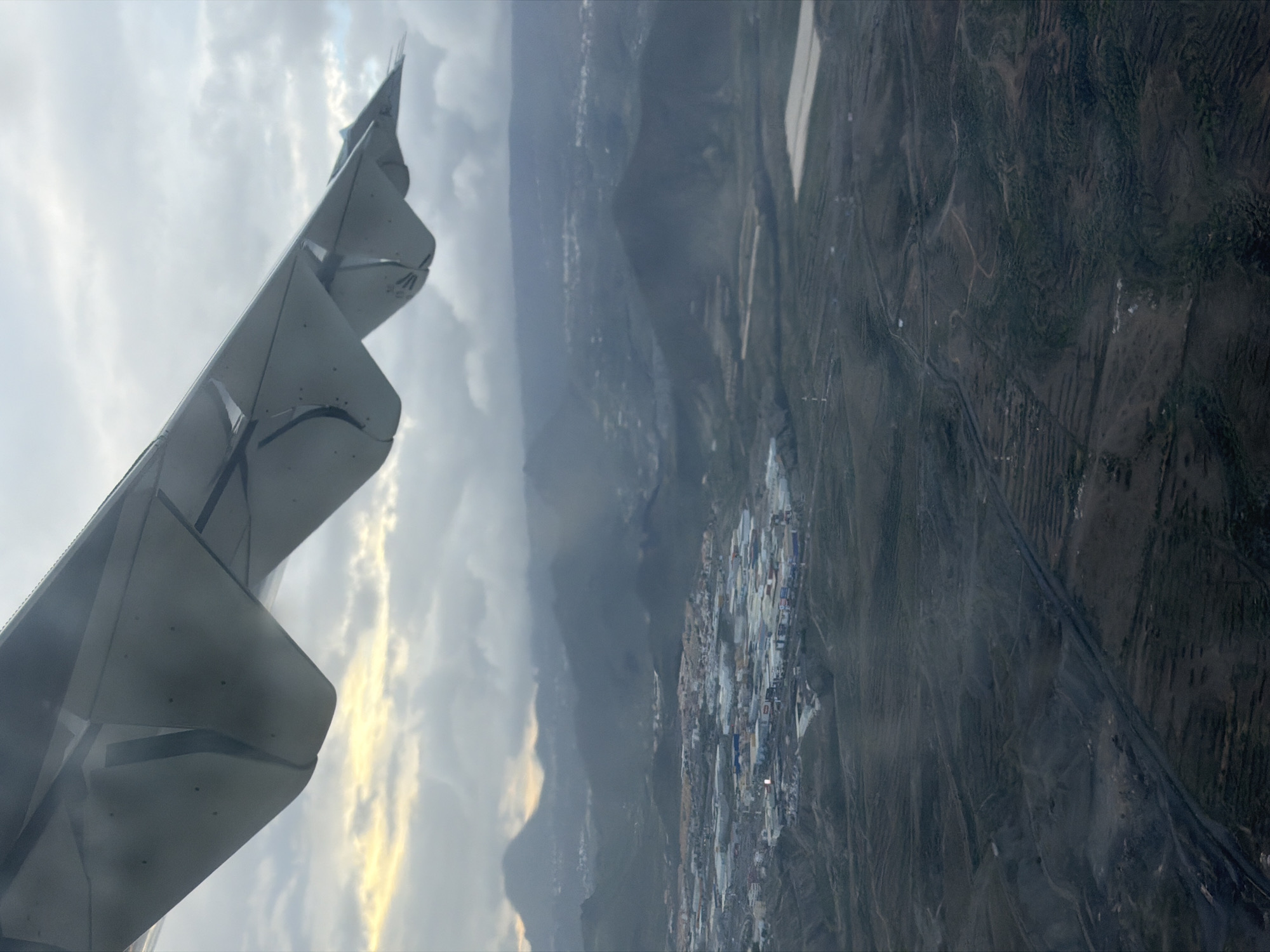 The image presents an aerial view captured from the window of an airplane, looking down at a landscape with a prominent aircraft wing in the foreground. The perspective is rotated 90 degrees counter-clockwise from a typical horizontal view, showing the wing on the left and the ground stretching to the right.
The left side of the frame is dominated by the light gray wing of the aircraft, featuring a distinct wingtip device (likely a sharklet or winglet) pointing upwards and slightly forward. Sections of the wing's trailing edge, possibly flaps or slats, are visible, suggesting the aircraft may be in a phase of flight like ascent or descent rather than cruise. A small, indistinct logo or text, possibly resembling a stylized "W" or "M," can be observed on the visible underside of the wingtip device.
Below and to the right of the wing, the diverse landscape of Granadilla de Abona, Spain, unfolds. In the midground, a significant urbanized or industrial area is visible, characterized by numerous light-colored, predominantly rectangular roofs, likely warehouses, factories, or commercial buildings. These structures, along with some darker roads or ground, suggest a modern development, potentially an industrial estate or port facility consistent with the Port of Granadilla. Some artificial lighting appears to be active within this complex, casting reflections or providing illumination.
Beyond this developed area, the terrain transitions into a more natural, arid, and hilly or mountainous landscape. Faint lines and patterns suggest roads, tracks, or terraced agricultural plots winding through the dark, rough ground. The overall weather appears partly cloudy or overcast, with patches of brighter, yellowish light breaking through the clouds on the horizon, hinting at either early morning or late afternoon. The scene conveys a sense of observation from above, capturing the interplay between natural landforms and human development under diffused light.