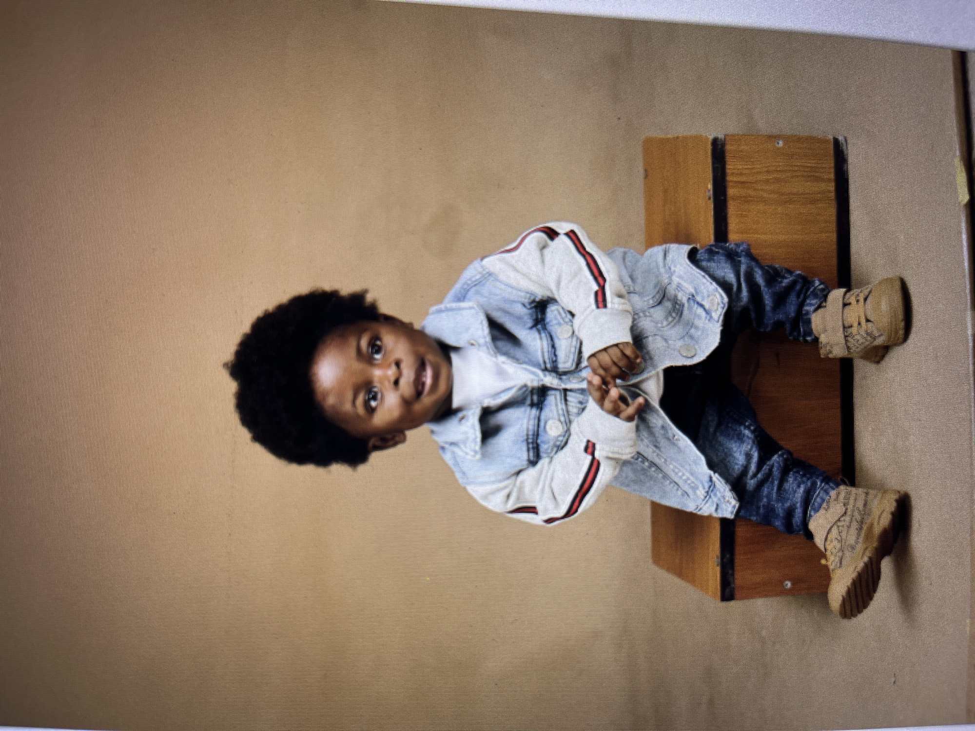 A young Black child with dark, curly hair is seated on a wooden box against a plain, tan background. The child is wearing a light blue denim jacket with gray sleeves that have red and black stripes, a white t-shirt, dark blue jeans, and tan sneakers with Velcro straps. The child is looking slightly to their left with a happy expression and their hands are clasped in front of them. The lighting is soft and even, creating a studio portrait feel. The image does not provide any information about the specific location in Gura Top, Nigeria.