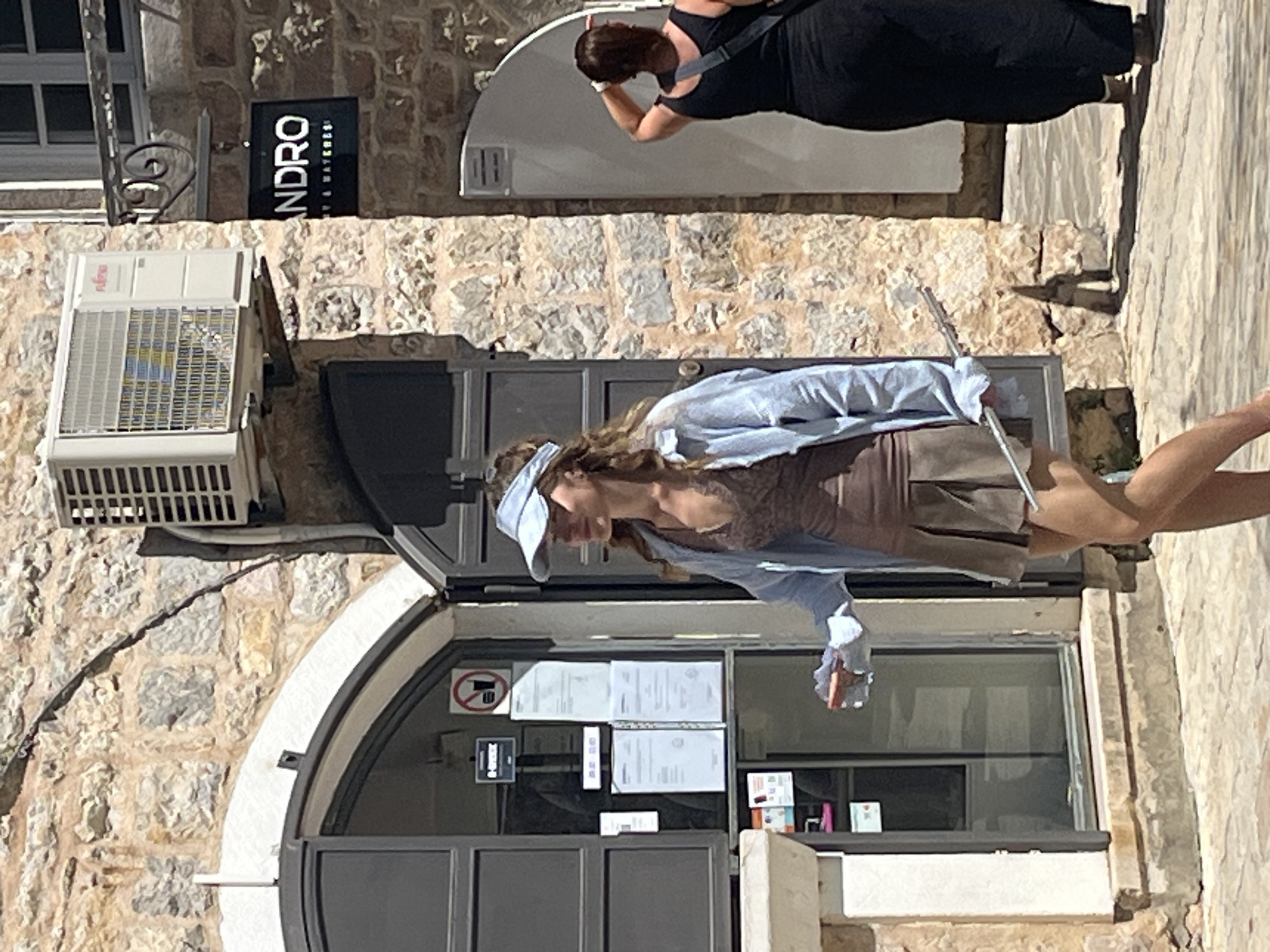 The image shows an outdoor scene in the city of Budva, Montenegro. The setting appears to be along a stone-paved street bordered by buildings constructed with light-colored stone. A young woman wearing a light blue visor, a brown dress with lace details, and a blue shirt is standing, seemingly holding something in her hand. A second woman, dressed in a black dress, stands in the background, near a white door. A sign that says "ANDRO" is visible on a nearby building. An air conditioning unit is affixed to the side of one of the stone buildings. A business is located within an arched doorway. Notices are posted on the interior of the glass windows.