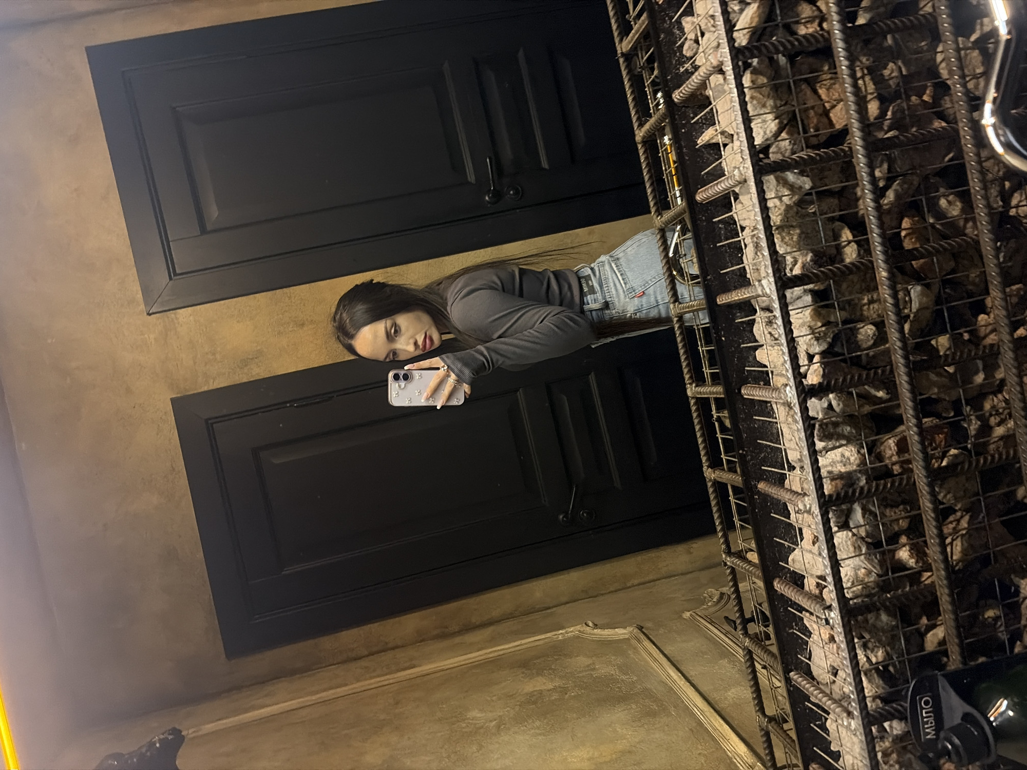 A woman is taking a mirror selfie indoors with a cell phone. The reflection shows two dark doors with dark handles and a stone wall behind the woman. Based on the "МЫЛО" (soap) label visible in the reflection and the architecture, the location is likely in Saint Petersburg, Russia.