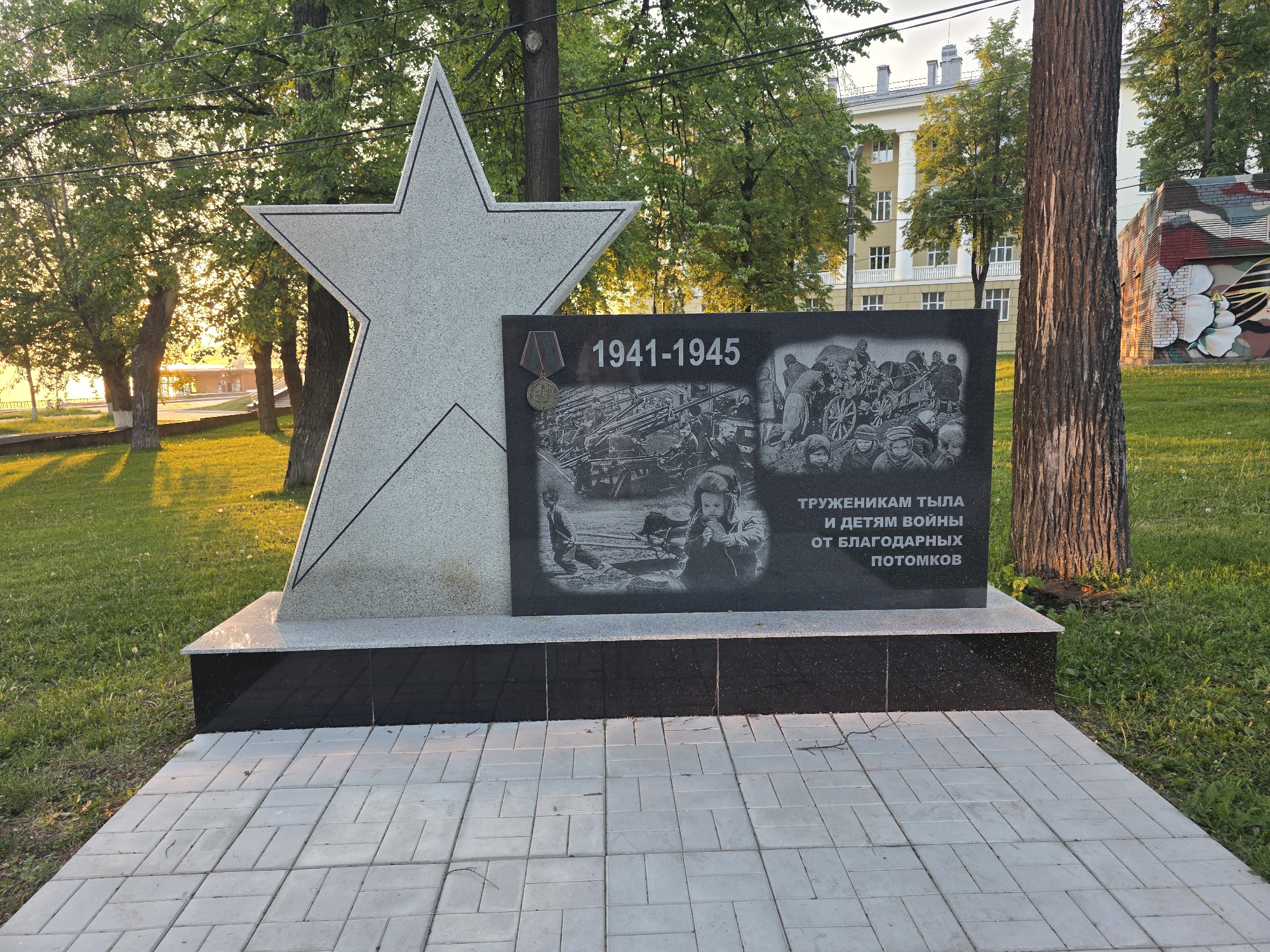 The image shows a monument dedicated to the workers of the home front and children of war in Votkinsk, Russia. The monument consists of a large gray star set upon a black base. Adjacent to the star is a dark gray plaque featuring the years 1941-1945 and images depicting wartime scenes of workers and children contributing to the war effort. The plaque also includes the inscription, "To the toilers of the rear and children of the war from grateful descendants" in Russian. The monument is situated on a paved area, with grass and trees in the background. In the background, there are buildings, one a yellow brick building and another brick structure bearing a mural. The scene appears to be outdoors in a park-like setting.