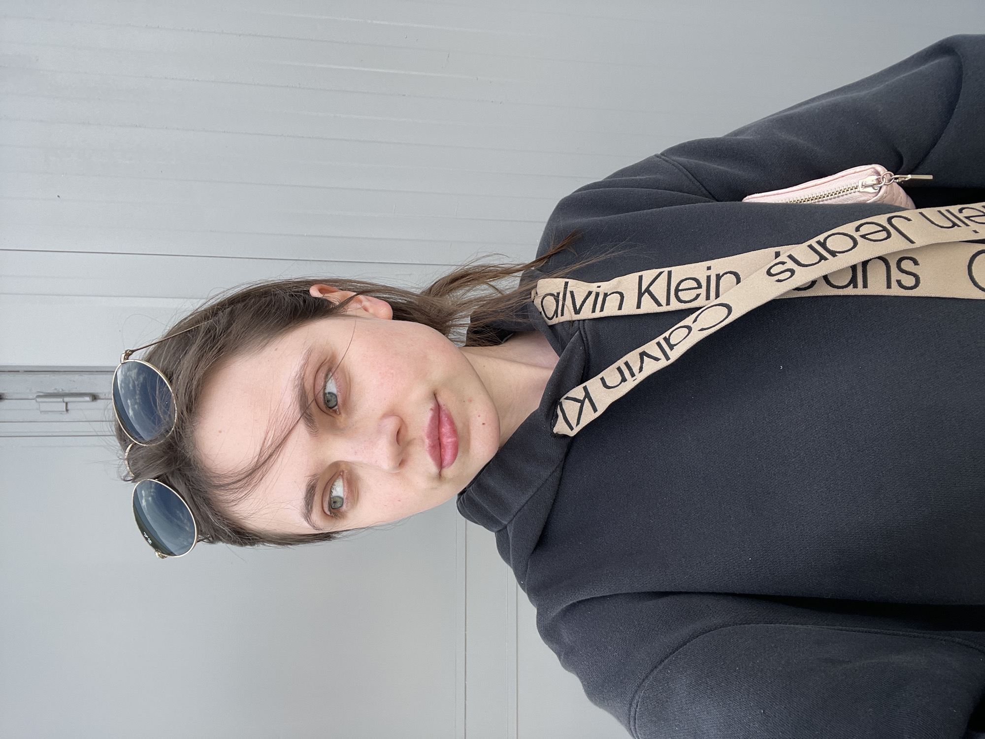 A woman poses for a selfie. She is wearing round sunglasses in her hair, a black hoodie with a Calvin Klein branded strap. She is in front of a light gray wall. The location is undetermined.