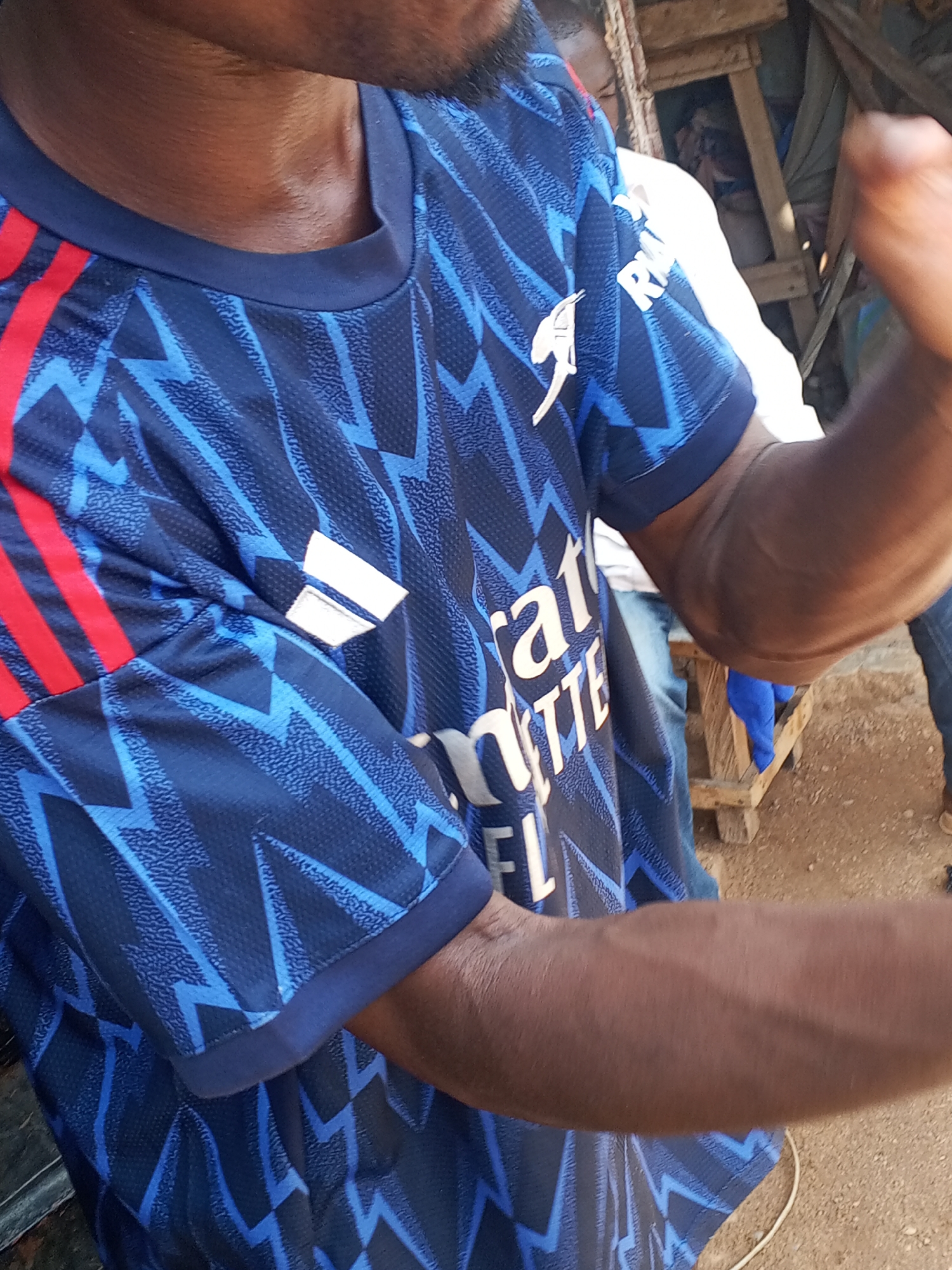 A person is wearing a blue Arsenal soccer jersey with red stripes on the sleeves and the Emirates logo. The location is an outdoor area with a wooden structure in the background and dirt ground. The person is flexing their arm. Another person is visible in the background wearing a white shirt and jeans.