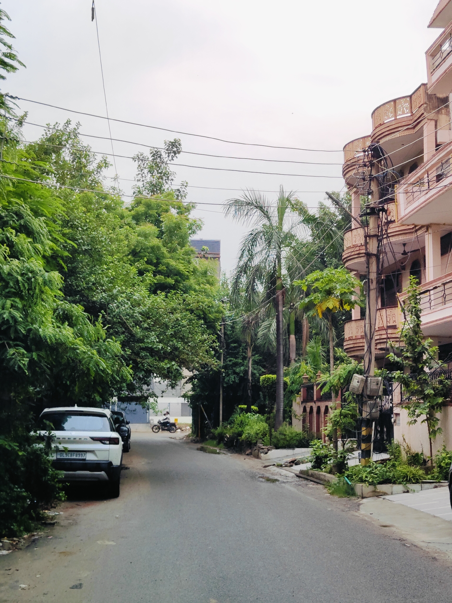 The image depicts a street scene in Gurgaon, India. On the left side of the street, a white SUV is parked, partially obscured by lush green foliage. Further down the road, a person is riding a motorcycle. The right side of the street features a multi-story building with balconies, supported by a utility pole laden with wires and electrical equipment. The sky is overcast. The road surface appears to be asphalt. Various trees and plants are visible throughout the scene.