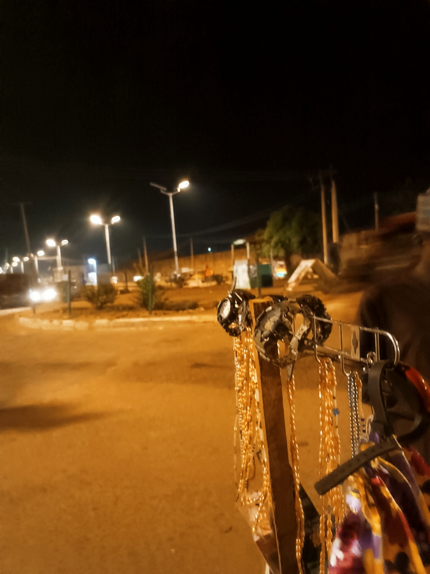 A nighttime street scene in Bukuru, Nigeria, featuring a roadside vendor's display. The vendor's wares consist primarily of gold-colored beaded necklaces and other accessories hanging from a metal and wood stand. The items appear to be jewelry and possibly fabric accessories. The street is illuminated by streetlights. A vehicle with its headlights on is visible in the distance. A person, presumably the vendor, is partially visible on the right side of the image. The background includes buildings, vegetation, and an elevated wall. The ground is paved and illuminated with a golden hue from the streetlights.