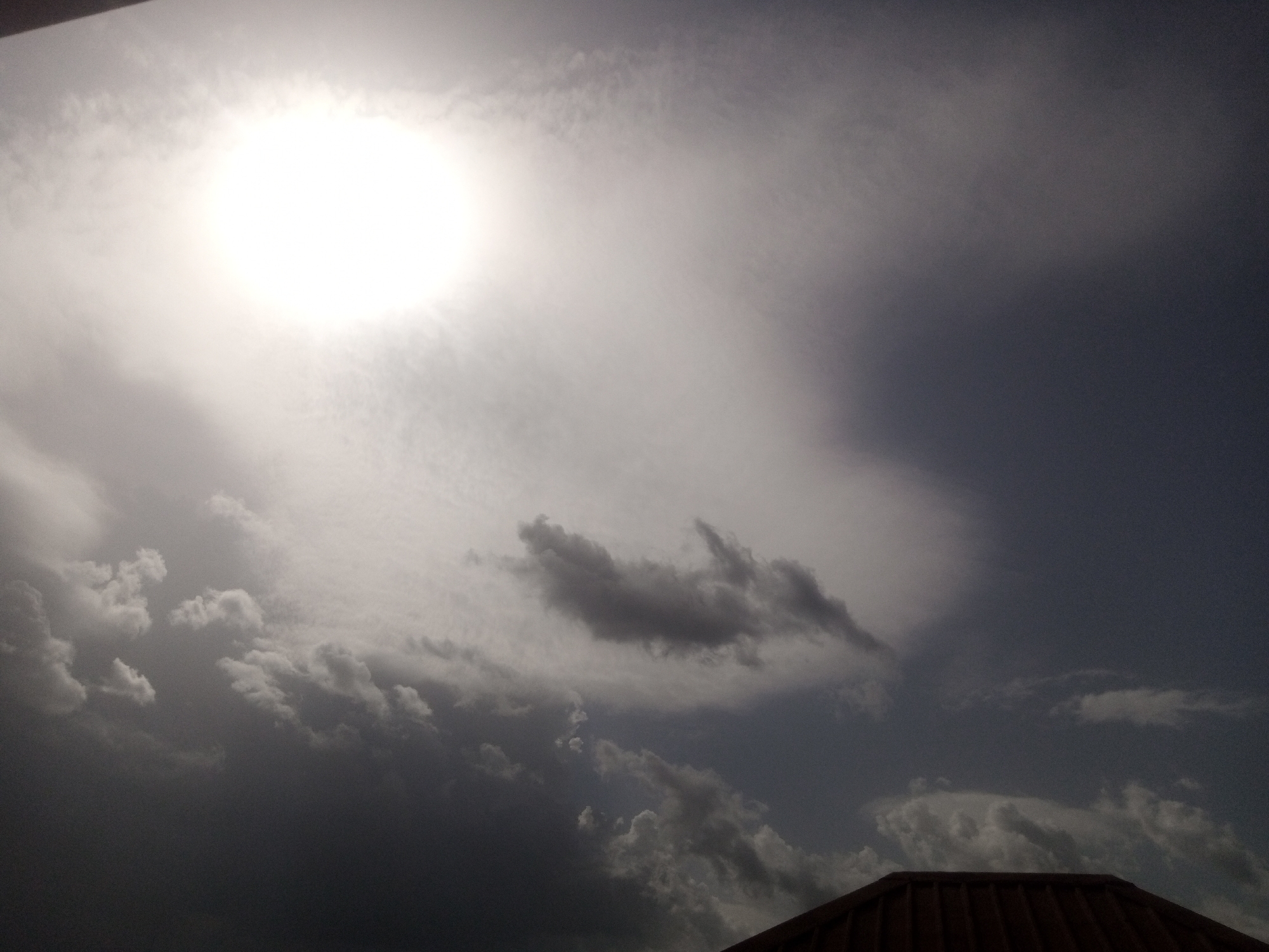 The image depicts a wide shot of a cloudy sky over a rooftop. The sun is visible as a bright, white disc in the upper left portion of the frame, partially obscured by thin, wispy clouds. The sky transitions from a bright, hazy area around the sun to darker, more ominous storm clouds towards the right and bottom of the image. The clouds are varied in texture and density, with some appearing thick and dark, while others are lighter and more diffused. In the lower right corner, the edge of a brown, corrugated rooftop is visible. The overall impression is of a dramatic sky, possibly before or during a storm, with the sun attempting to break through. No people or specific urban landmarks are discernible in the image.