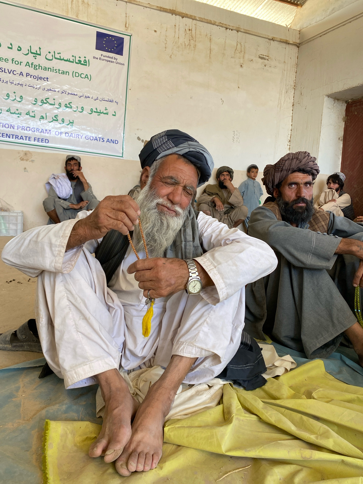 The image depicts a group of Afghan men and a boy seated in a room with a textured, light-colored wall and a corrugated metal ceiling. In the foreground, an elderly man with a long white beard, wearing a white shalwar kameez and a grey turban, is holding a string of prayer beads. He is wearing a wristwatch on his left wrist. Beside him, another man with a dark beard and a brown turban, also in traditional clothing, is looking towards the camera. In the background, other men and a young boy are seated on the floor. Behind them, a banner with writing in both Pashto and English is displayed, indicating that the location is Pasab, Afghanistan, and that the event is related to a "SLVC-A Project" and a "CONCENTRATE FEED PROGRAM OF DAIRY GOATS AND". The room appears to be sparsely furnished, with some crates visible in the background. The lighting suggests it is daytime.