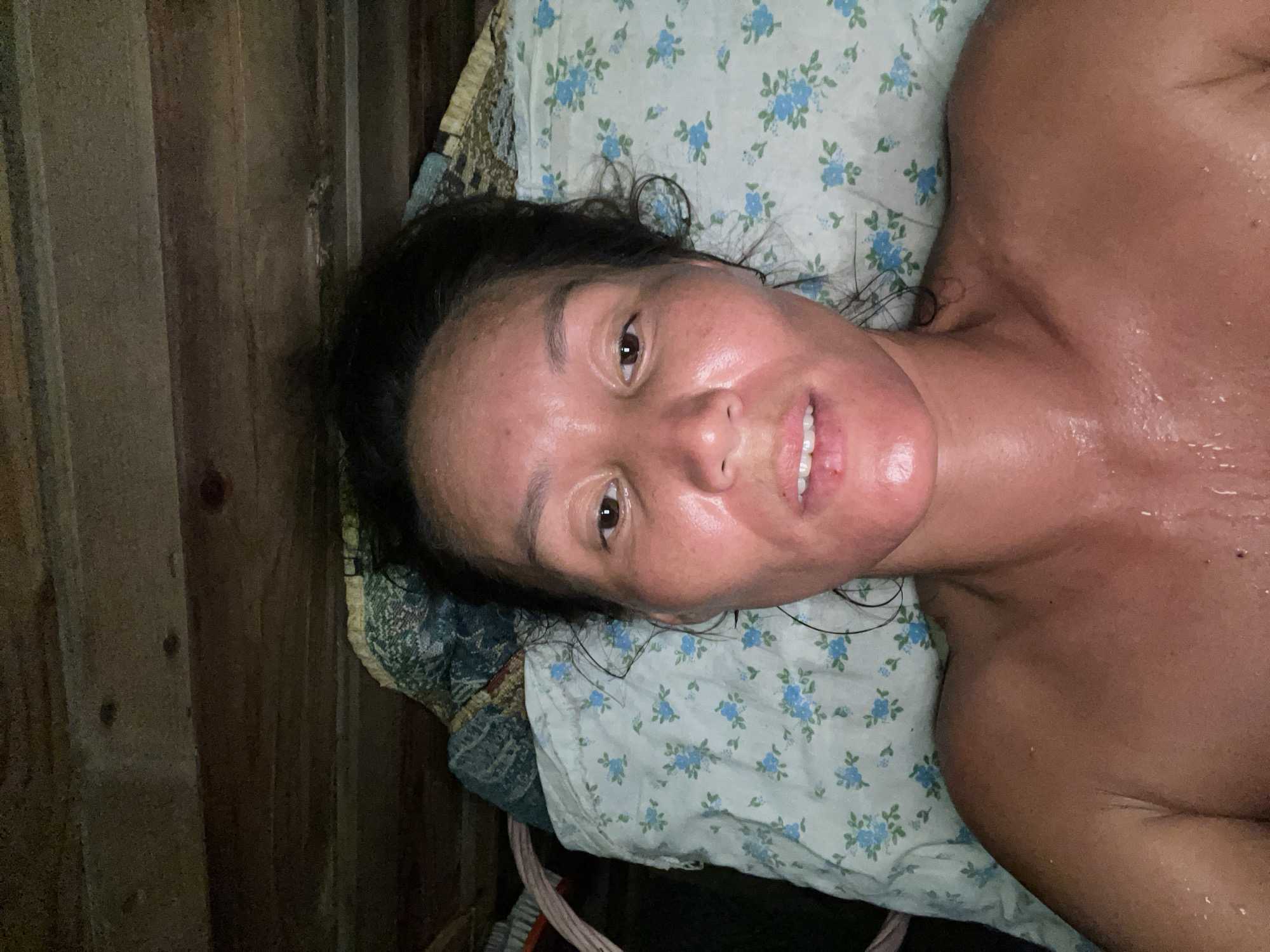 A woman is lying on a floral-patterned bedsheet in what appears to be a wooden room. Her skin is shiny and appears to be sweating, suggesting a warm environment. The lighting is dim, casting shadows on the wooden walls behind her. She is looking directly at the camera with her mouth slightly open, revealing her teeth. The image is oriented vertically, with the woman's head towards the right side of the frame.