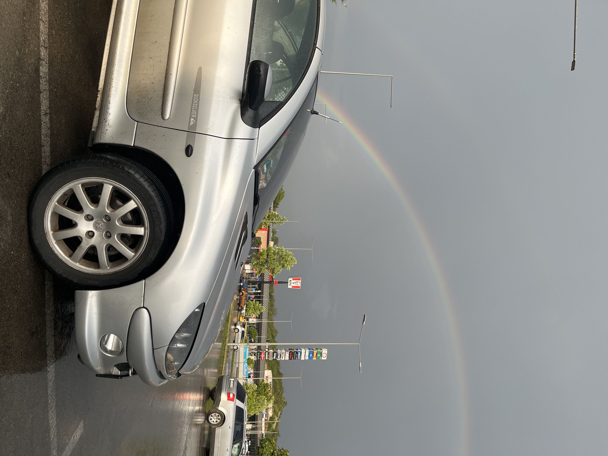 In Shumen, Bulgaria, a silver Peugeot 206 is parked in a wet parking lot under a cloudy sky where a double rainbow is visible. The car is the primary subject, showing its front passenger side and part of the driver's side, with water droplets evident on its surface. The "Quiksilver" logo is visible on the car's door. In the background, there are other vehicles, streetlights, trees, and commercial buildings including a KFC restaurant and a tall sign listing various businesses like Billa, Pepco, Douglas, and Deichmann. The ground is wet and reflective, indicating recent rain. The overall scene captures a moment after a rain shower in an urban commercial area.
