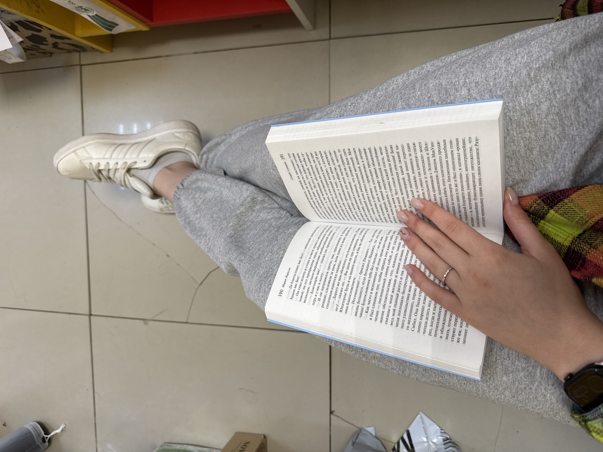 A person is seated, reading a book. They are wearing grey sweatpants and white sneakers. The person's hand is resting on the book, and a ring is visible on their finger. They are also wearing a smartwatch with a black band and a green, yellow, and black plaid strap. The book appears to be open to pages 190 and 191, with text in a foreign language. The background is a tiled floor and a red and yellow shelf. There are other items scattered on the floor.