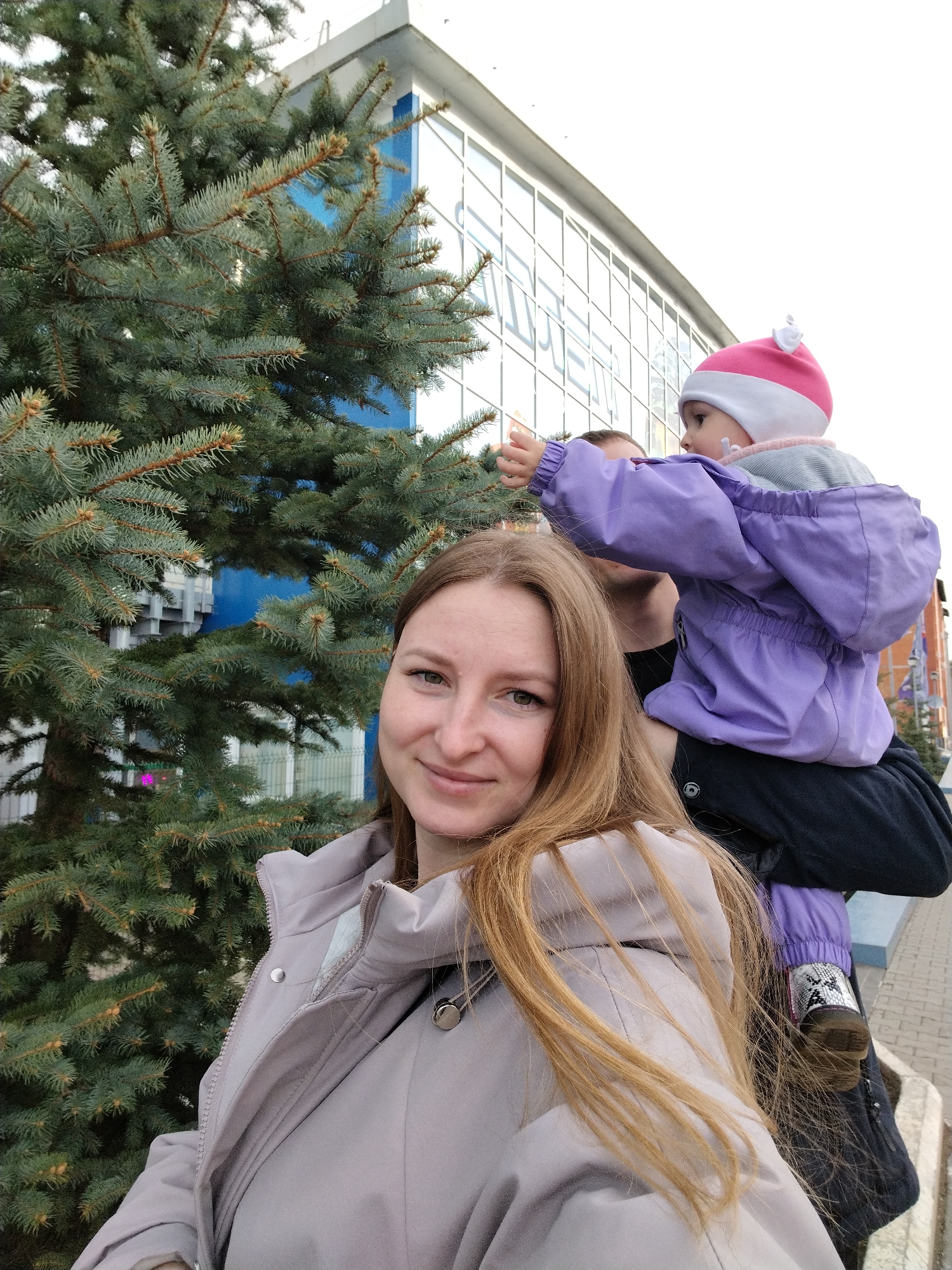 In Chaikovsky, Russia, a woman with long, light brown hair and wearing a light grey jacket smiles at the camera. Behind her, a man carries a child on his shoulders; the child is dressed in a purple coat and a pink-and-white hat, reaching out toward a blue spruce tree. The family stands on a brick-paved walkway in front of a building with a blue and white facade featuring many windows.
