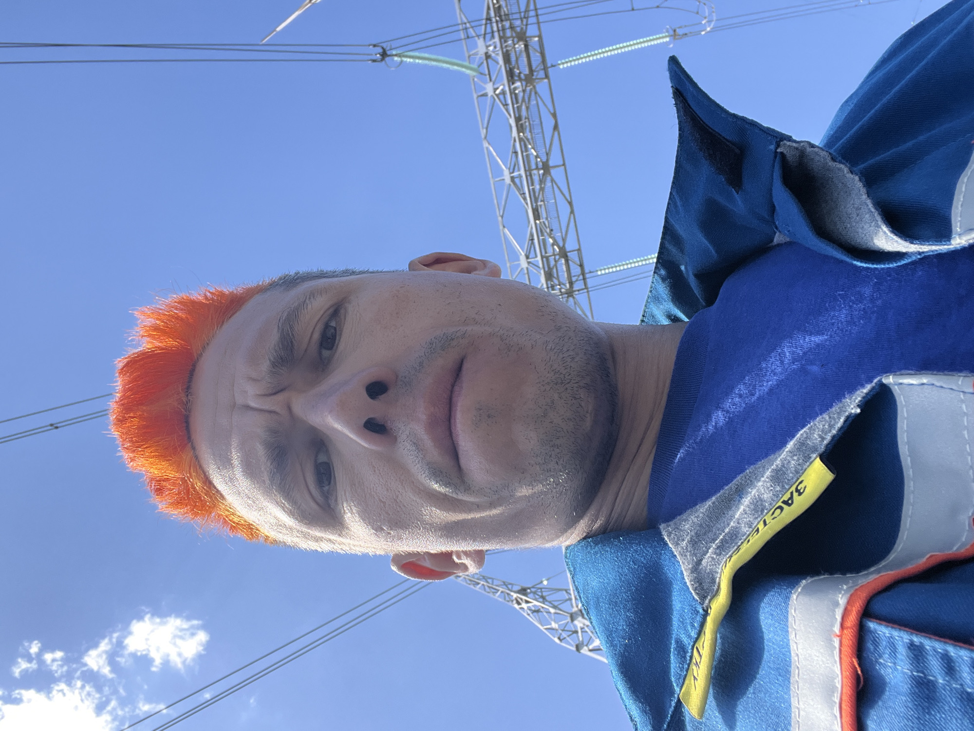 A close-up, low-angle selfie depicts a man with closely cropped, vibrant orange hair. He has fair skin, stubble, and a slightly furrowed brow. He is wearing a blue and grey work uniform with reflective strips and a yellow tag. Behind him is a bright blue sky with a few wispy clouds. Overhead power lines and a metal power tower structure are visible. The location is likely in or near Chaikovsky, Russia, given the typical infrastructure present.