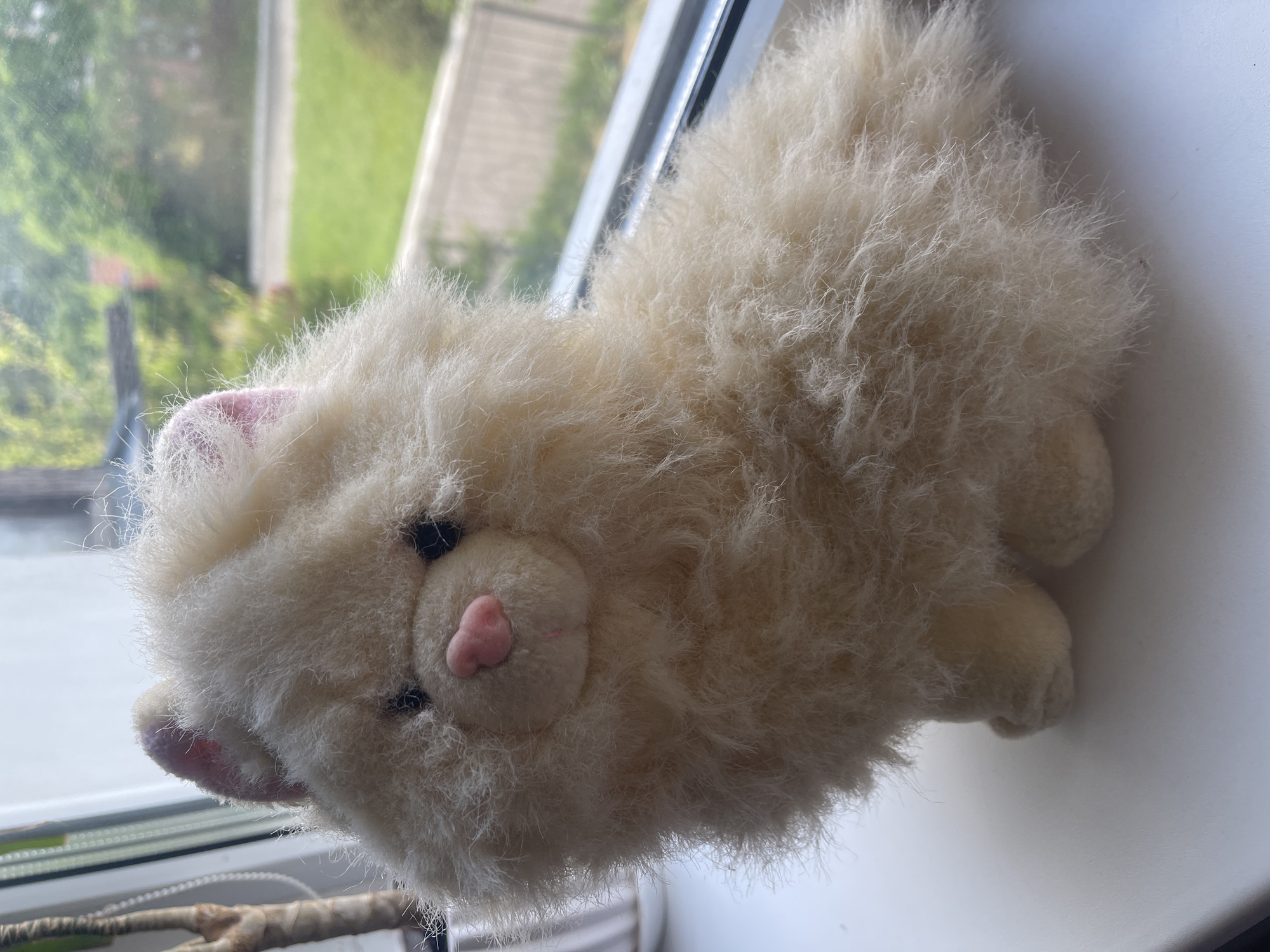 A cream-colored, fluffy stuffed cat toy sits on a white surface, likely a windowsill, with a blurred green outdoor scene visible through a window in the background. The cat has small black eyes, a pink nose, and pink inner ears. Its fur is long and wispy. To the left of the cat, a portion of a plant with a brown stem is visible. The lighting suggests it is daytime, with natural light coming from the window.