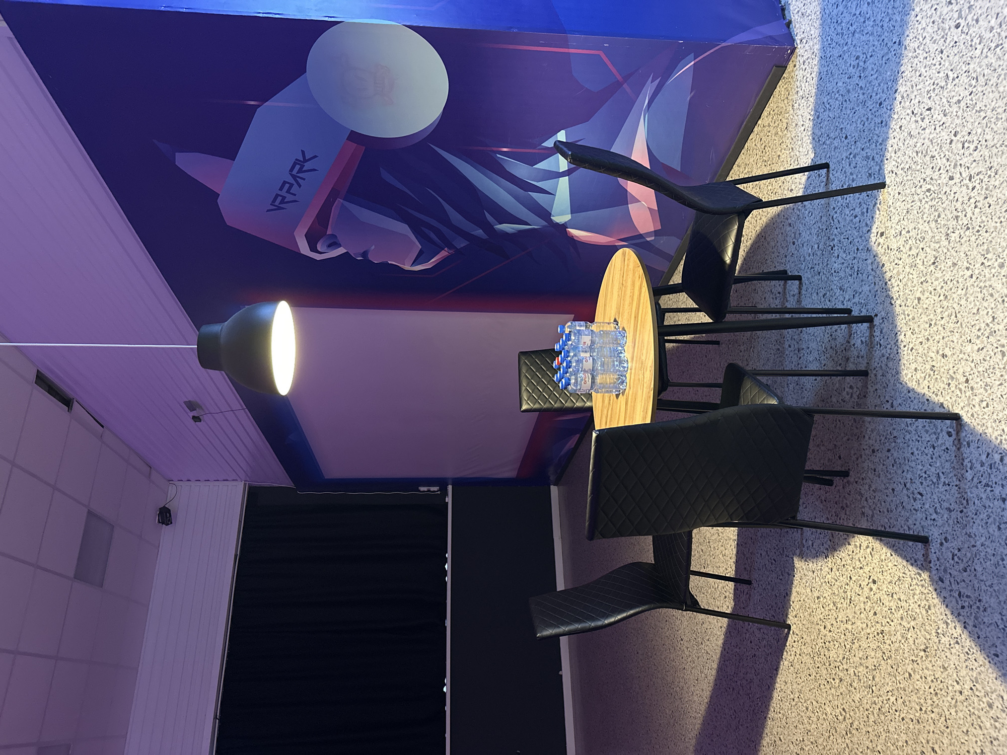 The image shows a modern indoor space with a round wooden table surrounded by black chairs. The scene is illuminated by a hanging lamp with a bright bulb. On the table are several bottles of water. The backdrop features a mural with the word "VR PARK" visible. The flooring appears to be a speckled, light-colored material. A dark partition is partially visible in the background, along with a tiled wall. The image provides no information about the location city or state.